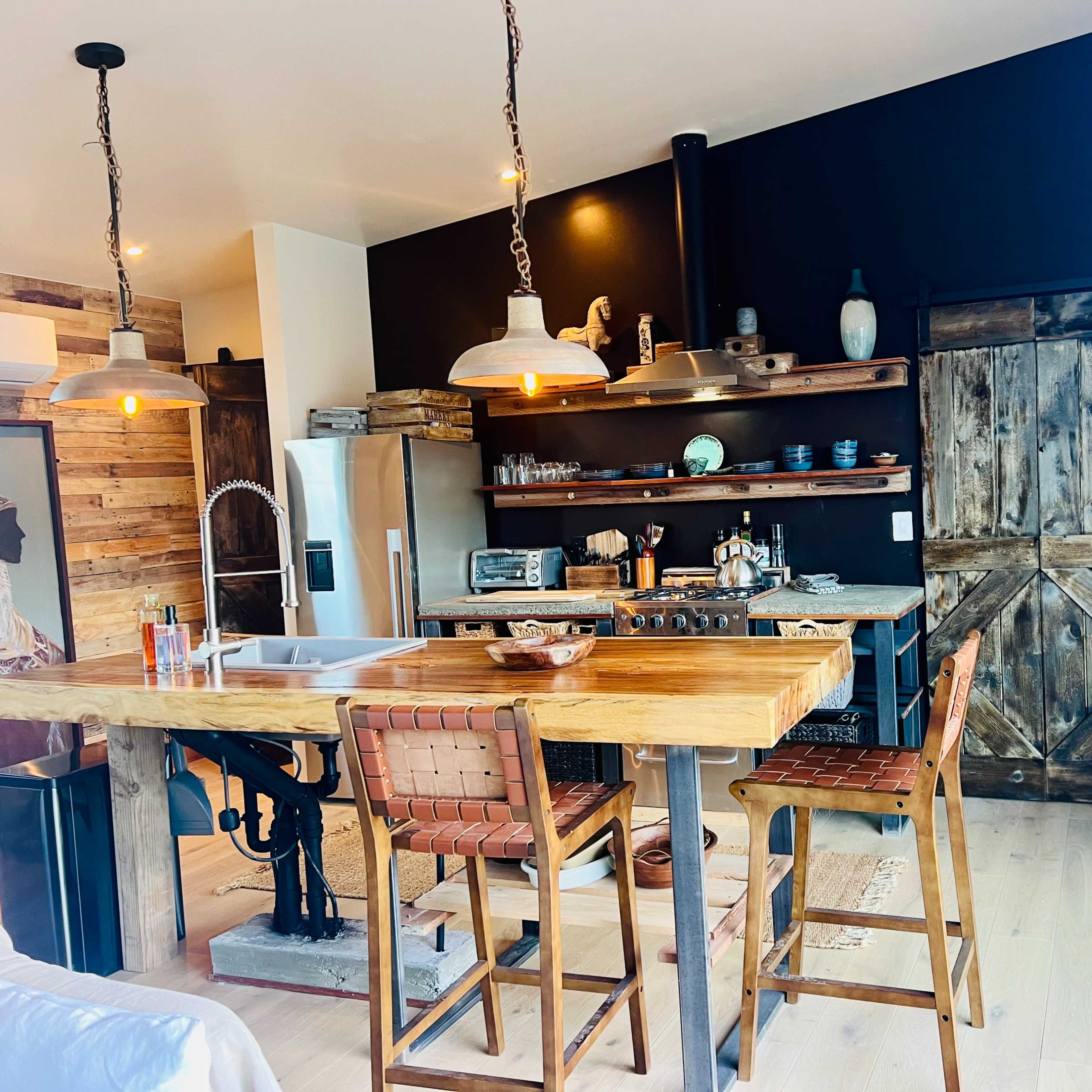The kitchen features a wooden dining table with metal bar stools, open shelves displaying dishware, and a rustic door leading to another space.