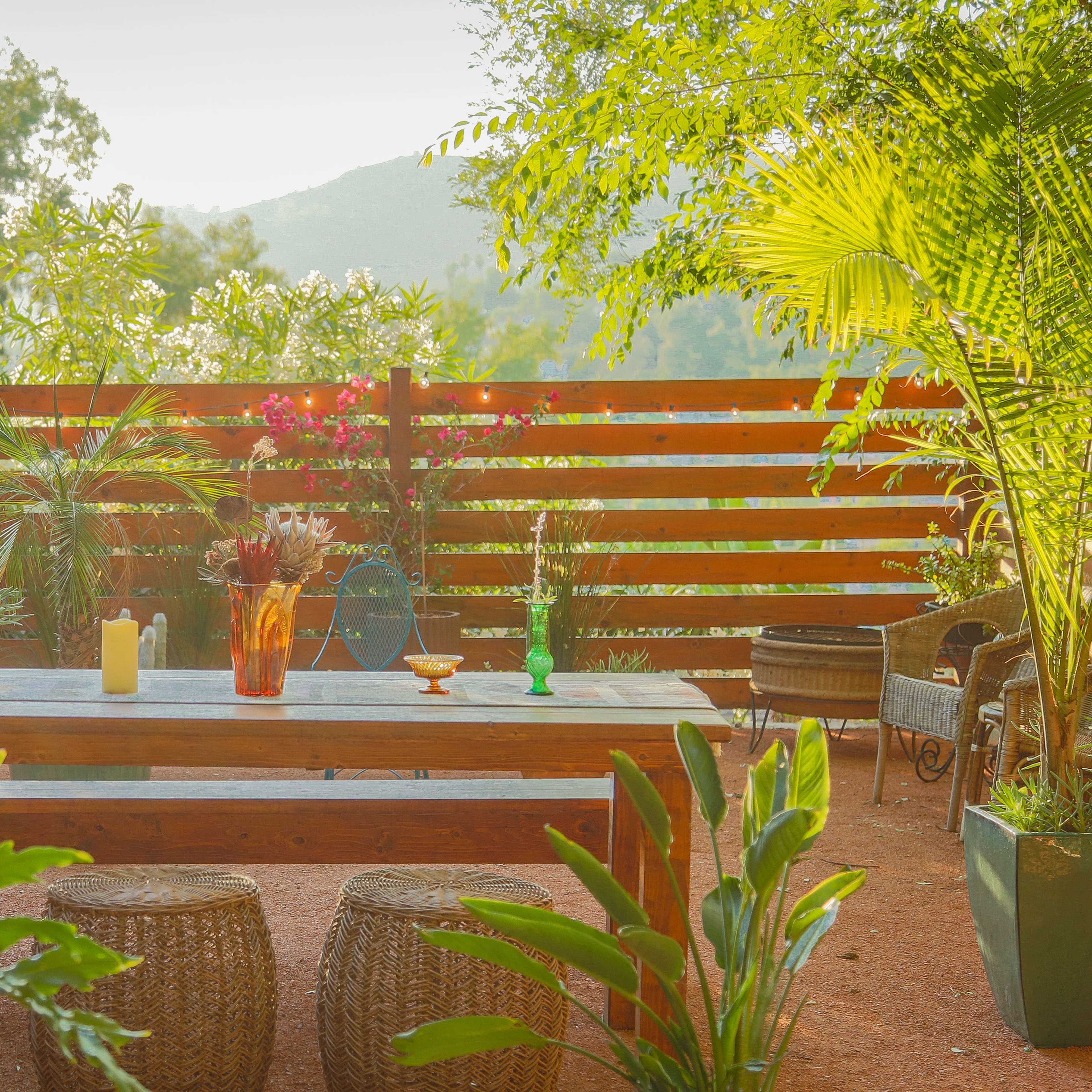A wooden table is set in a lush garden surrounded by potted plants and a wooden fence, with a view of distant hills.