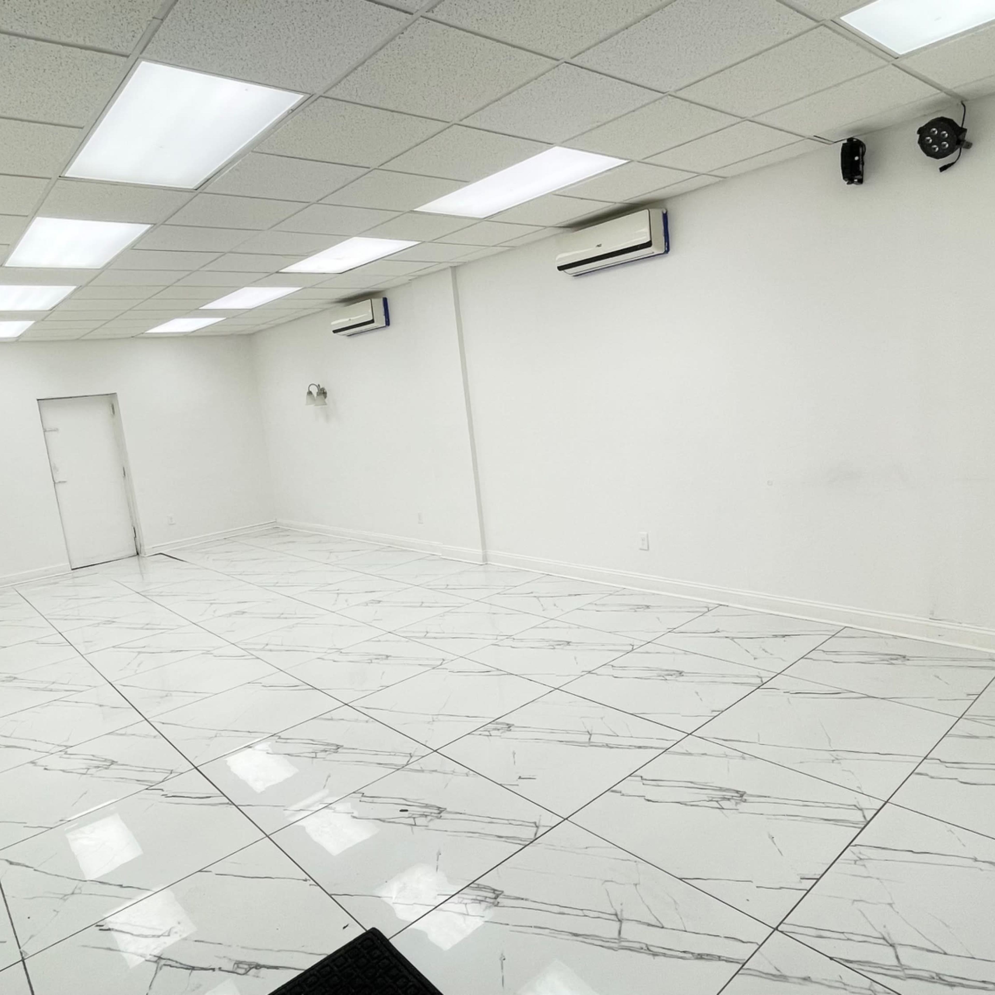 A spacious, empty room with white walls and a glossy marble-patterned floor, featuring ceiling lights and air conditioning units.
