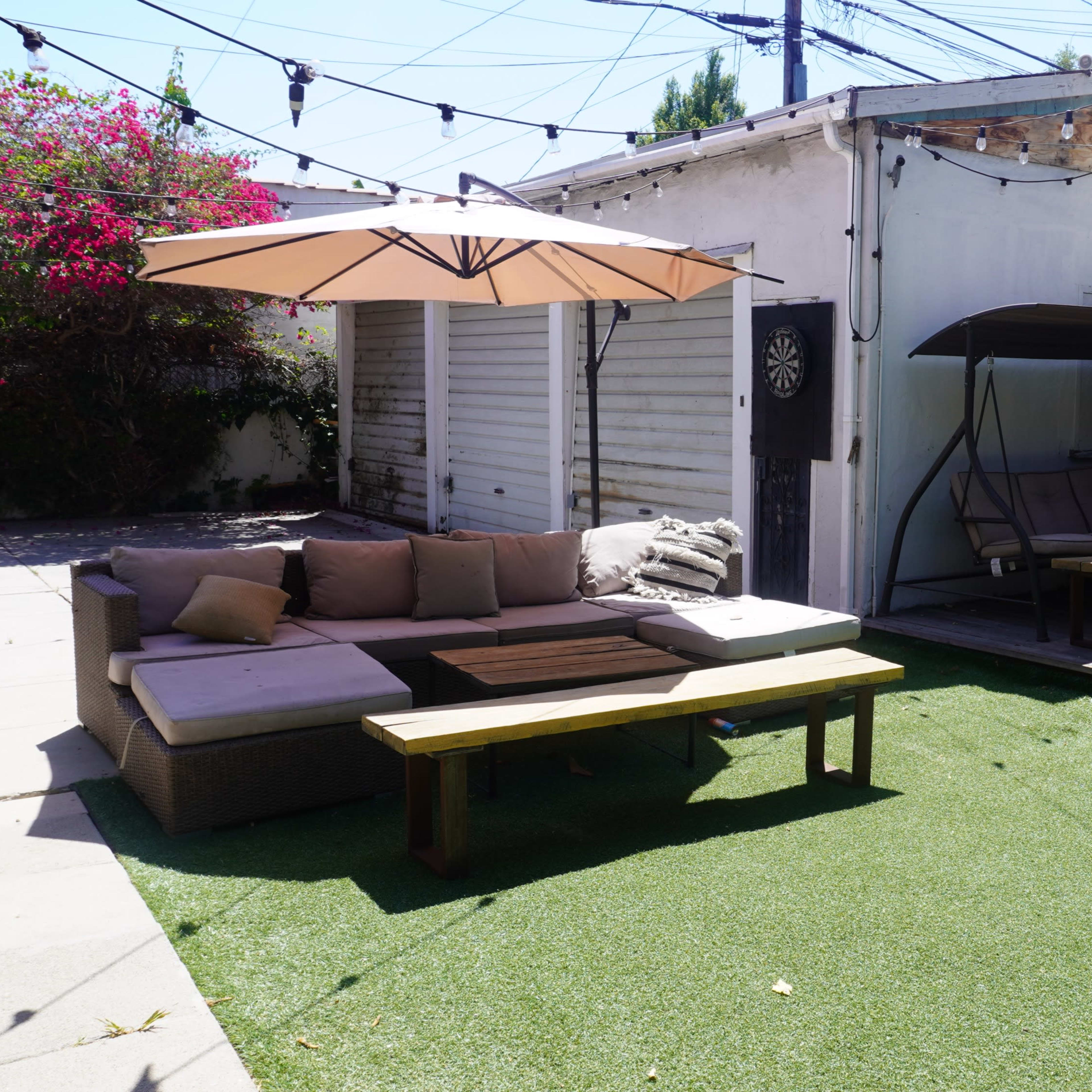 The scene features a patio area with a sectional sofa, a wooden coffee table, two shaded lounge areas, and artificial grass surrounded by vibrant plants and utility lines.