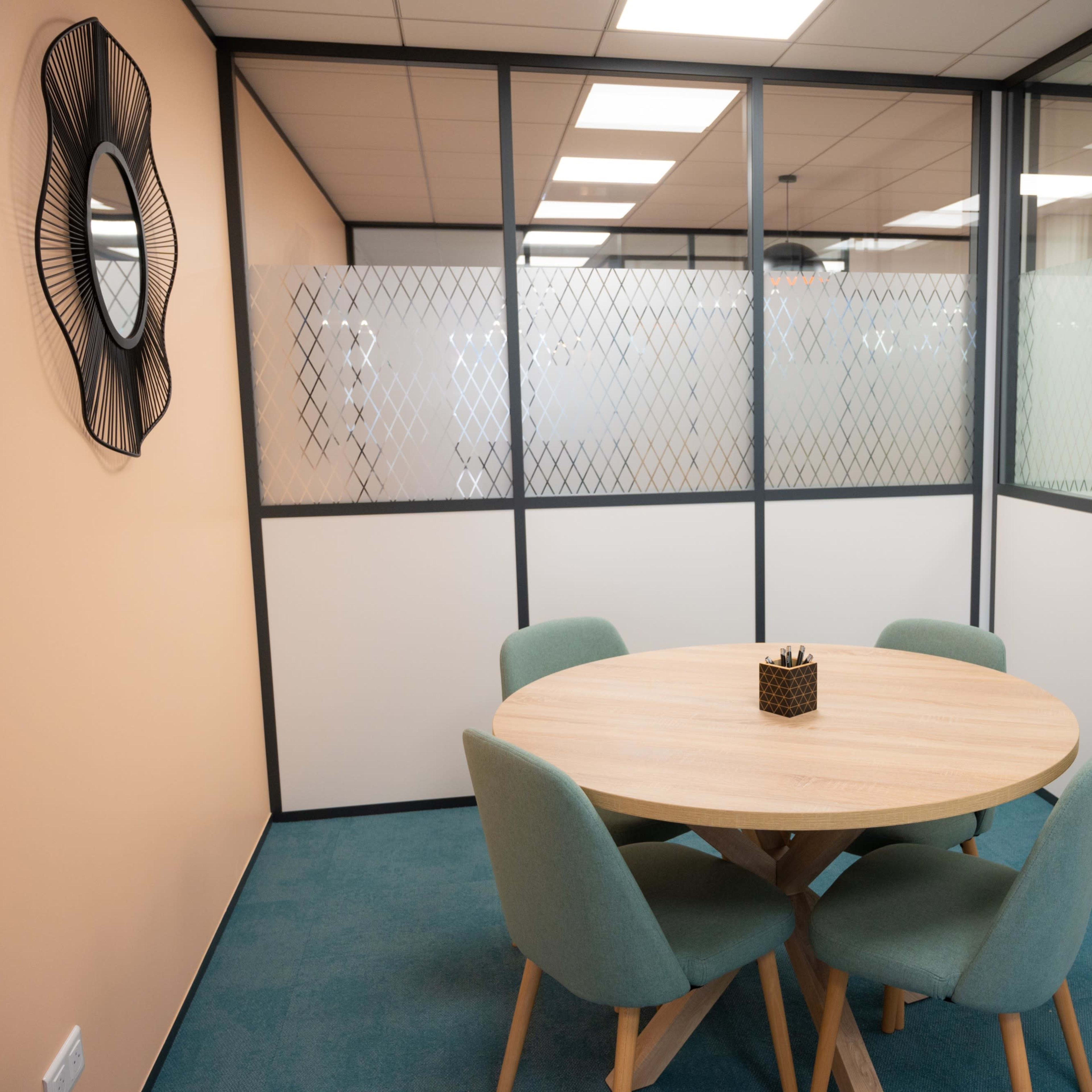 A modern meeting room with a round wooden table surrounded by four teal chairs, enclosed by glass walls.