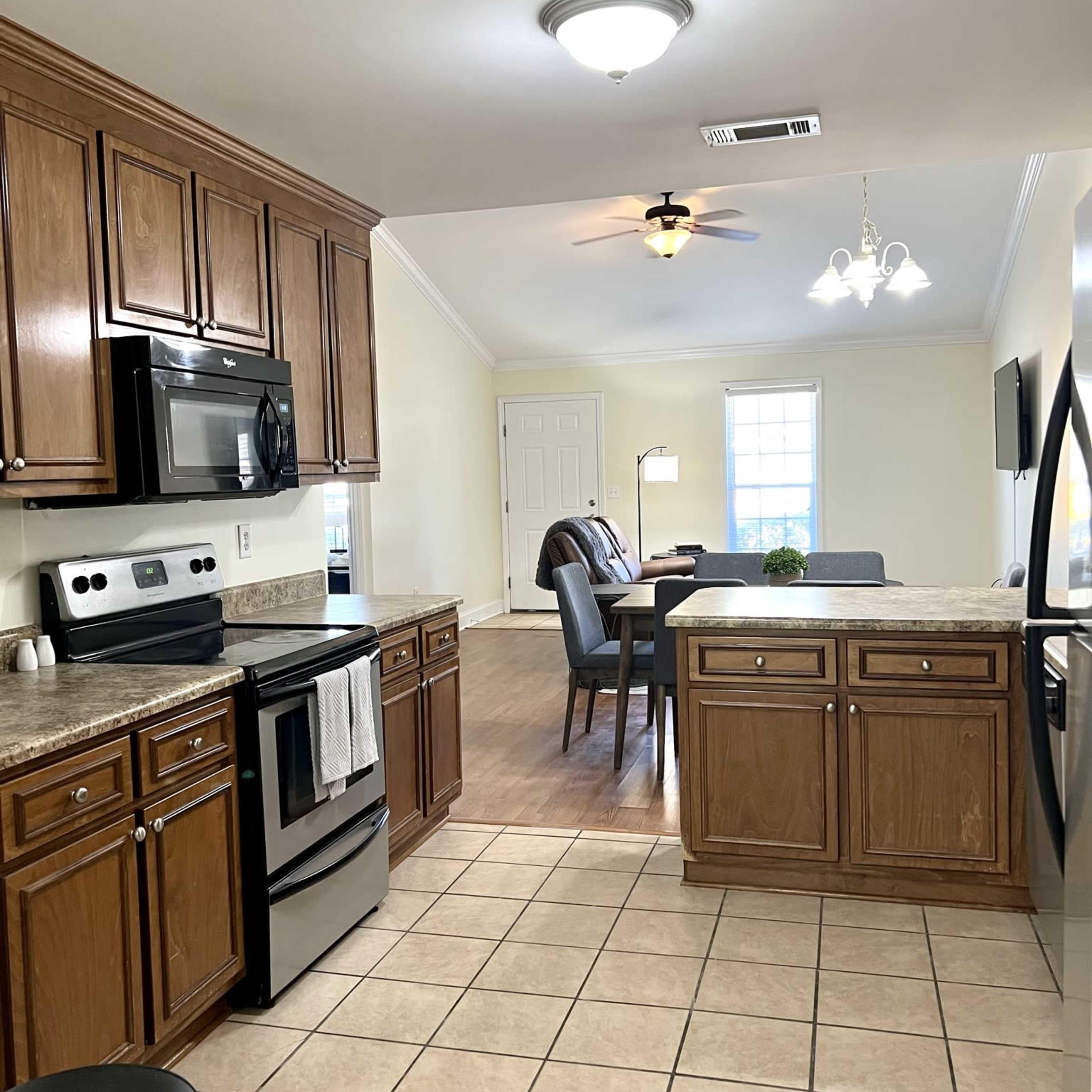 A modern kitchen features wooden cabinets, stainless steel appliances, and a tiled floor, with an adjoining living area visible in the background.