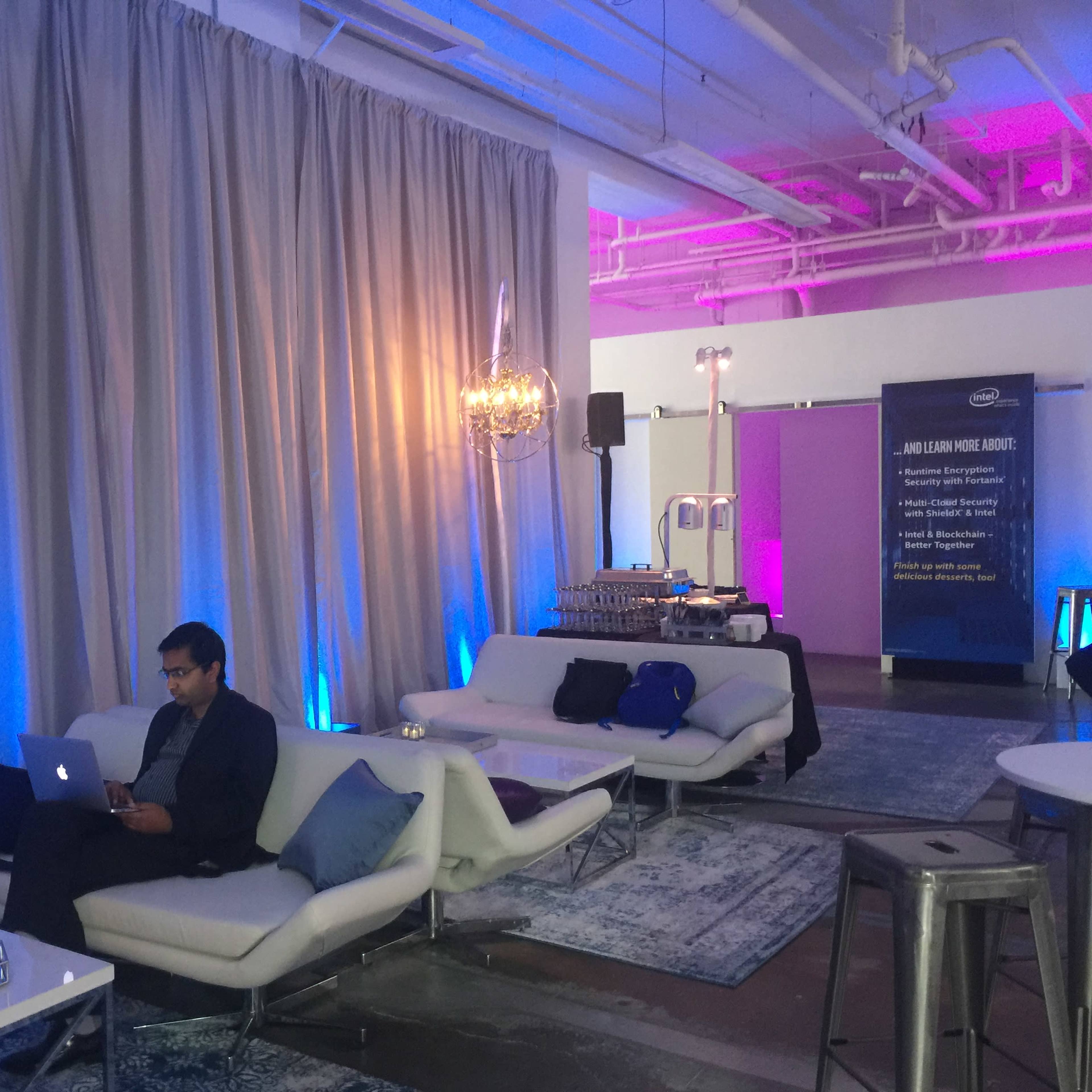 A man sits on a white couch using a laptop in a lounge area with purple and blue lighting, surrounded by draped curtains and tables with refreshments.