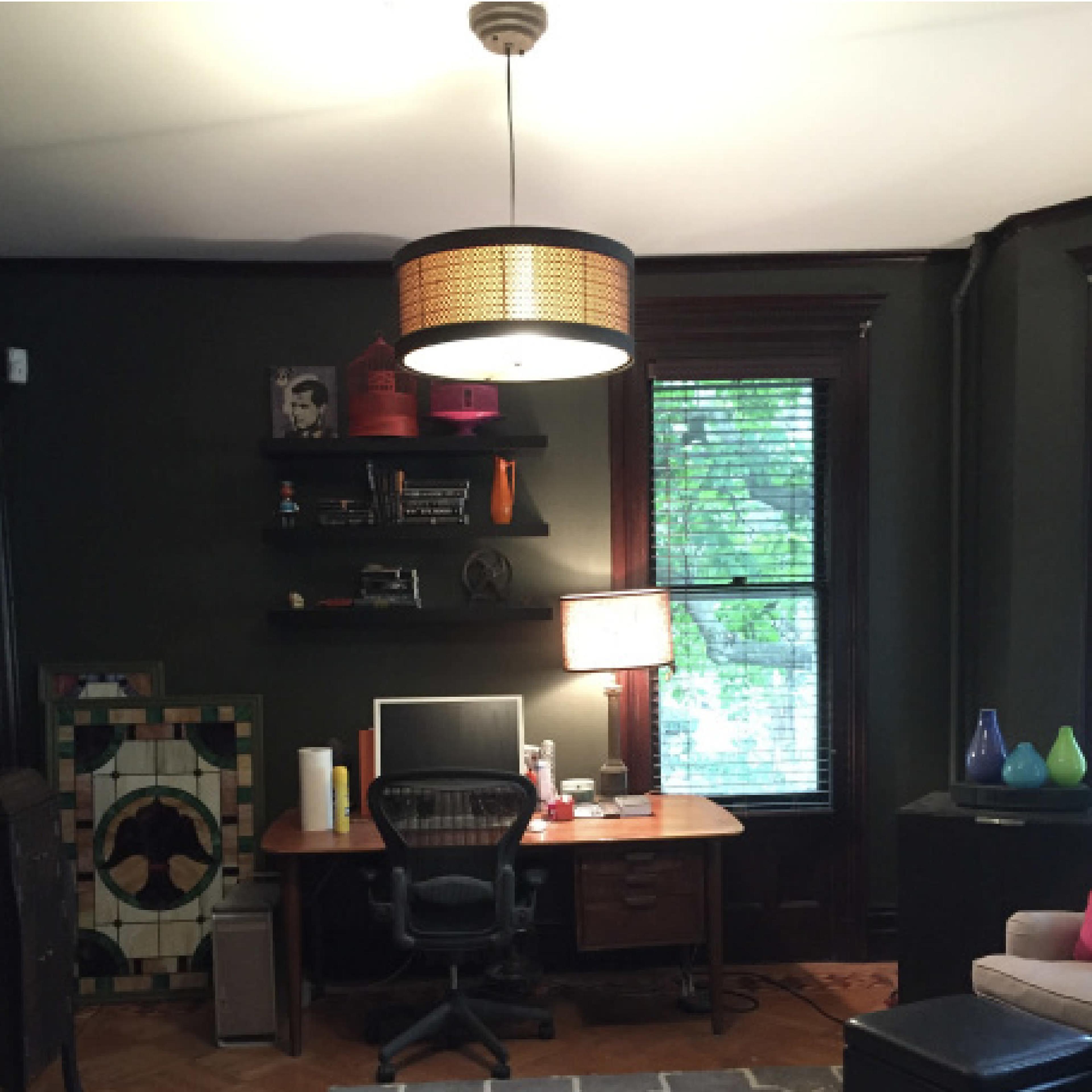 A room with a dark green wall, a wooden desk with a computer, a lamp, bookshelves, and a patterned rug.