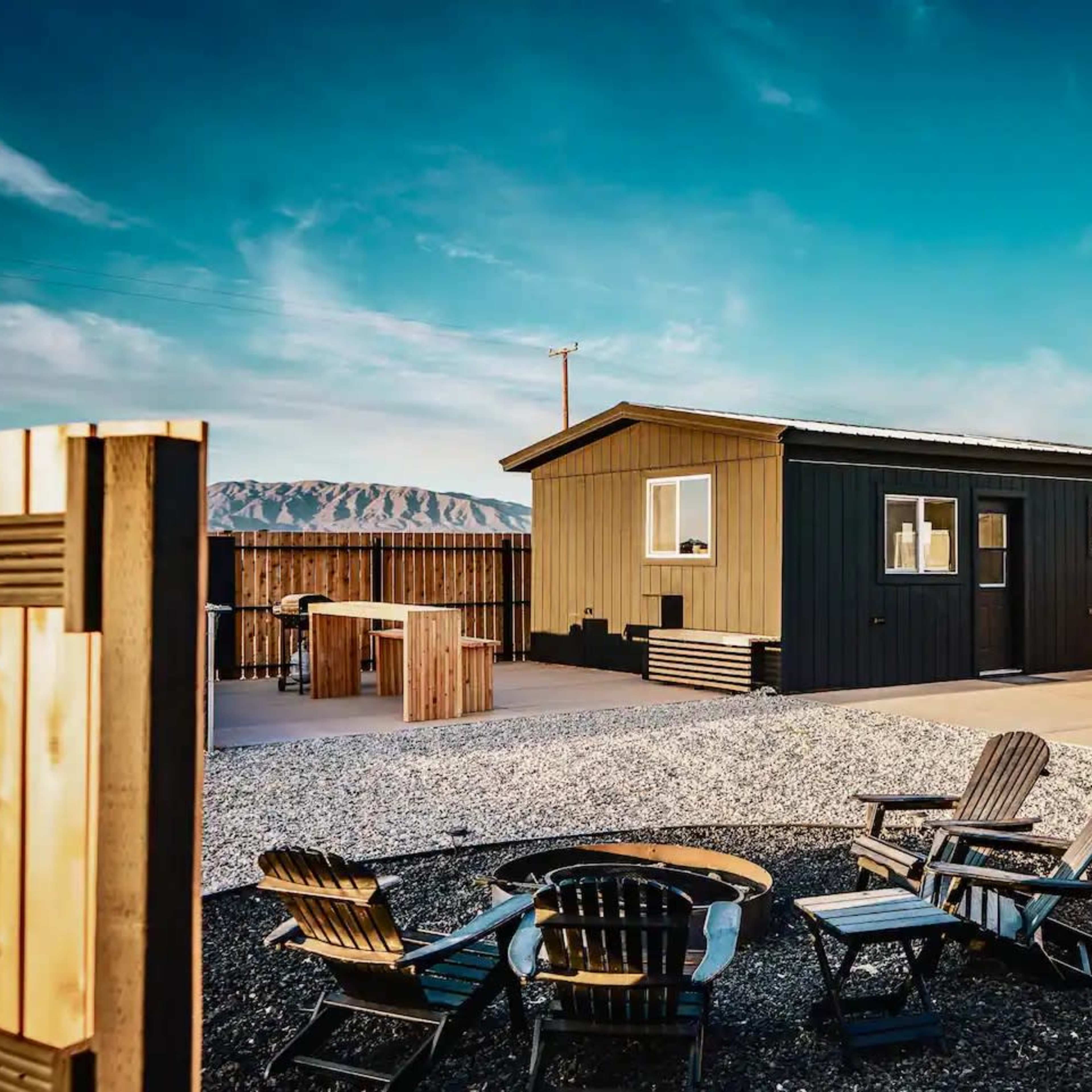 A modern black and yellow house with a gravel yard and a fire pit surrounded by wooden chairs.