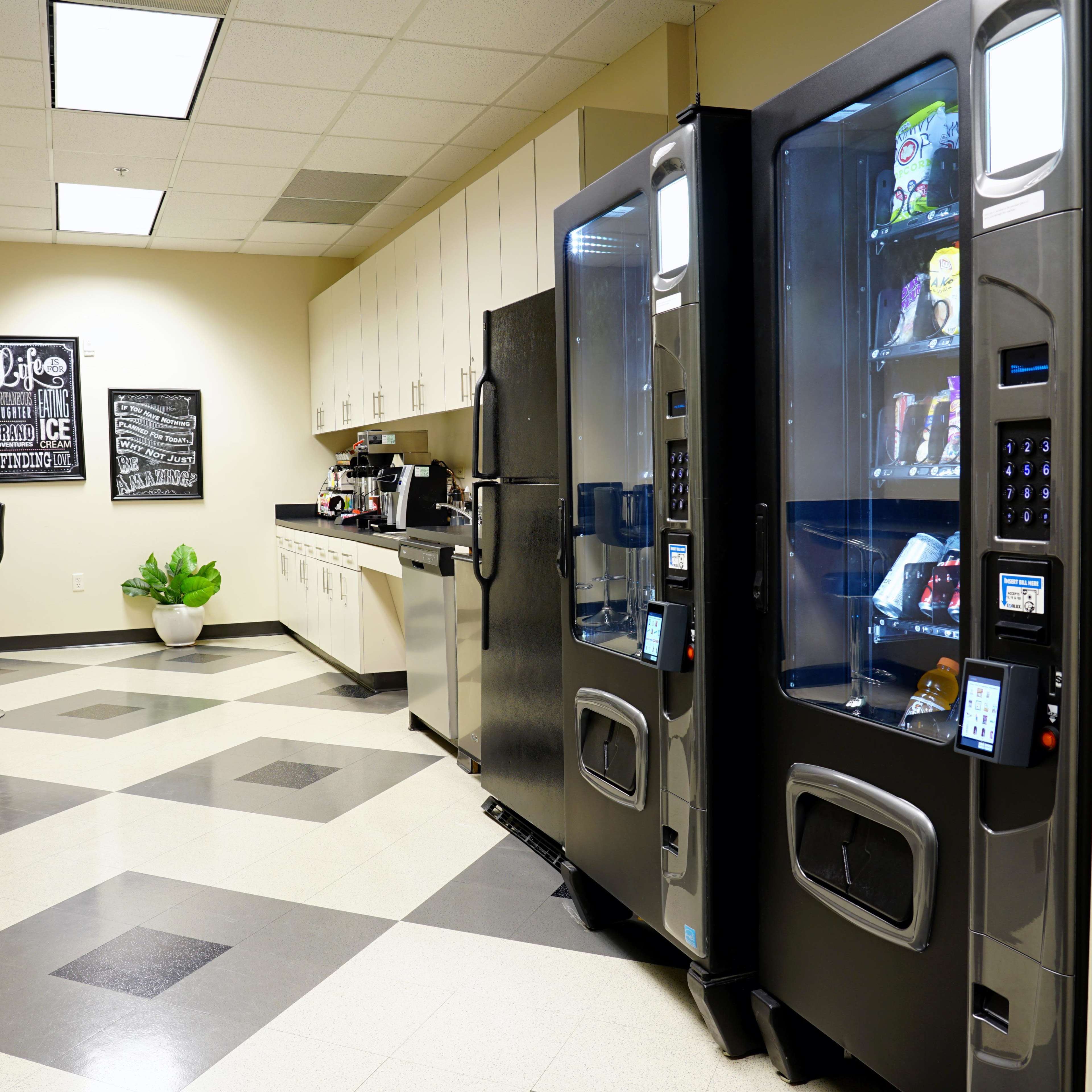 A break room features two vending machines alongside a kitchenette with cabinets, a refrigerator, and seating.