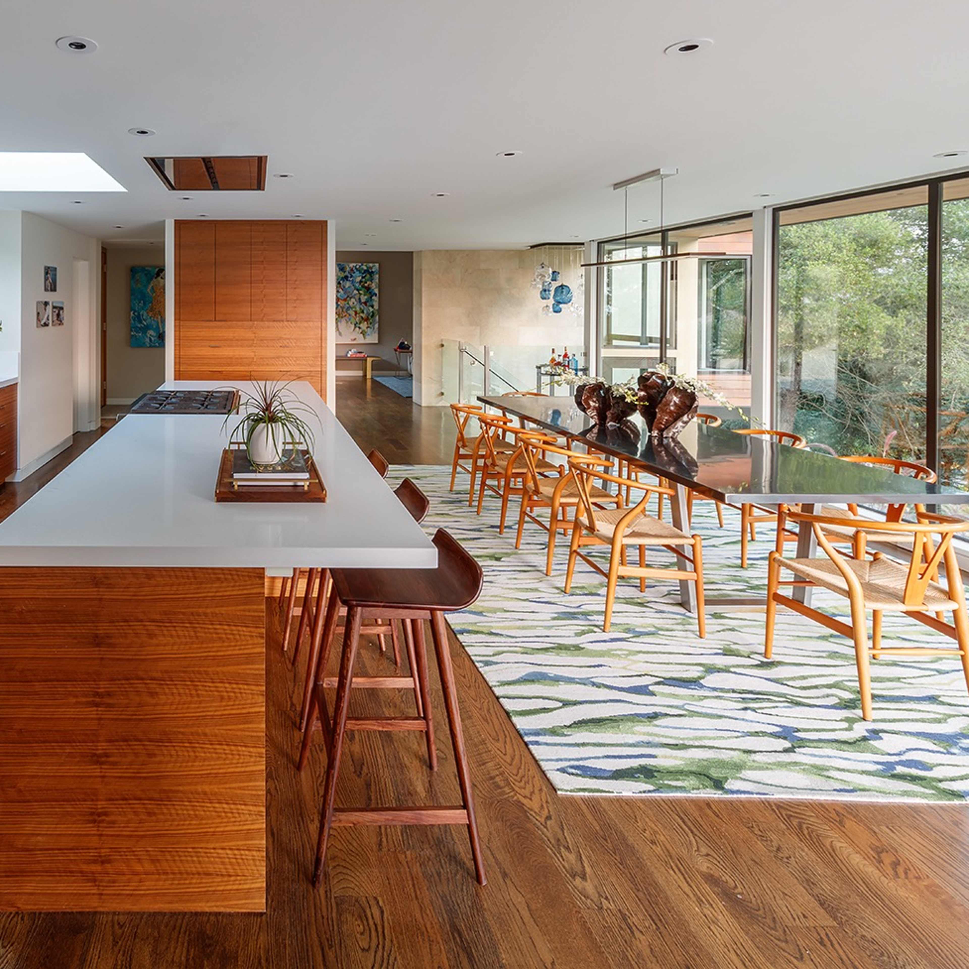 A modern kitchen features wooden cabinetry, a central island with bar stools, and a dining area lined with large windows overlooking a garden.