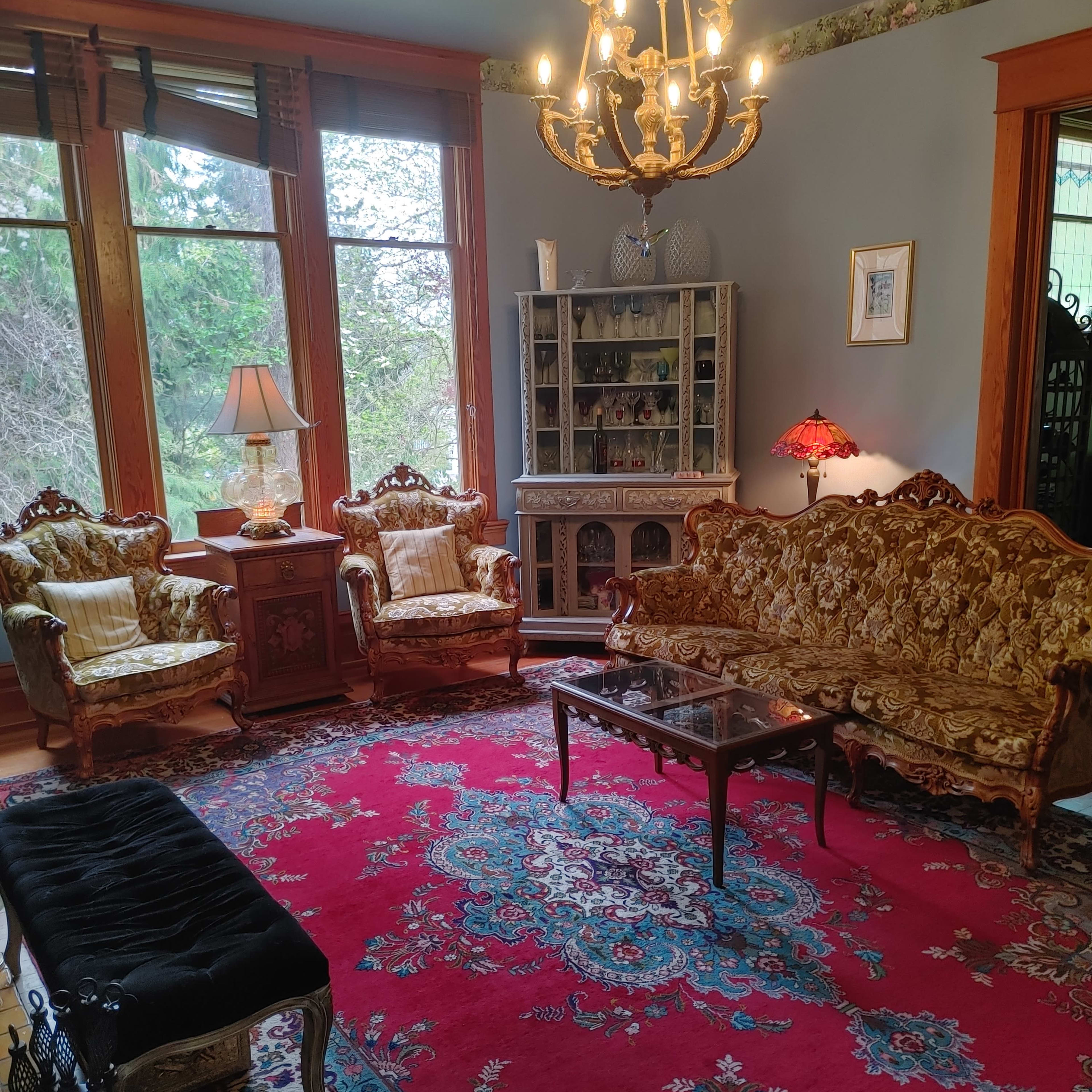 The room features ornate furniture, including two armchairs and a couch, surrounding a central coffee table on a large patterned rug, with a chandelier hanging from the ceiling.
