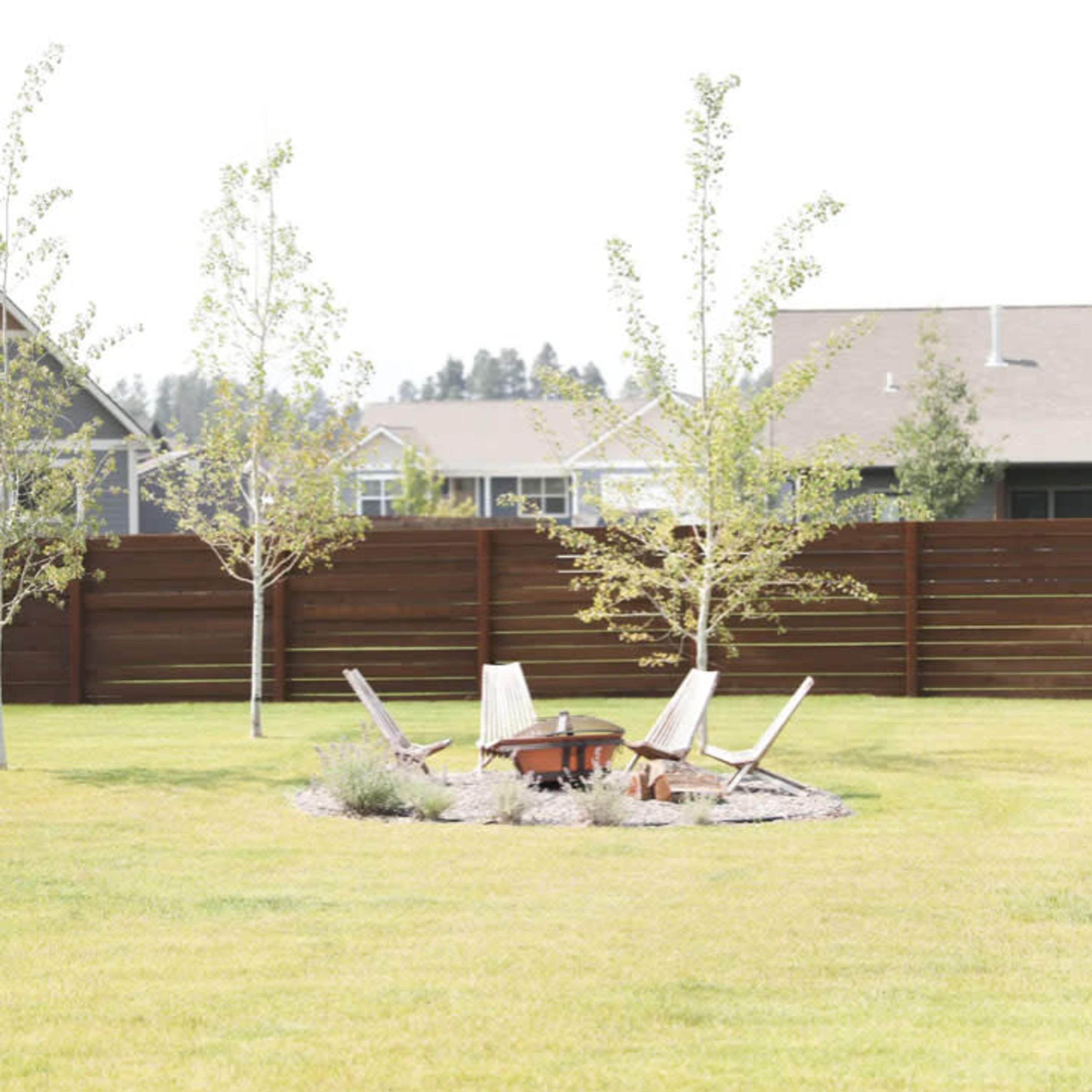A circular fire pit surrounded by four chairs on a grassy lawn, with small trees and a wooden fence in the background.
