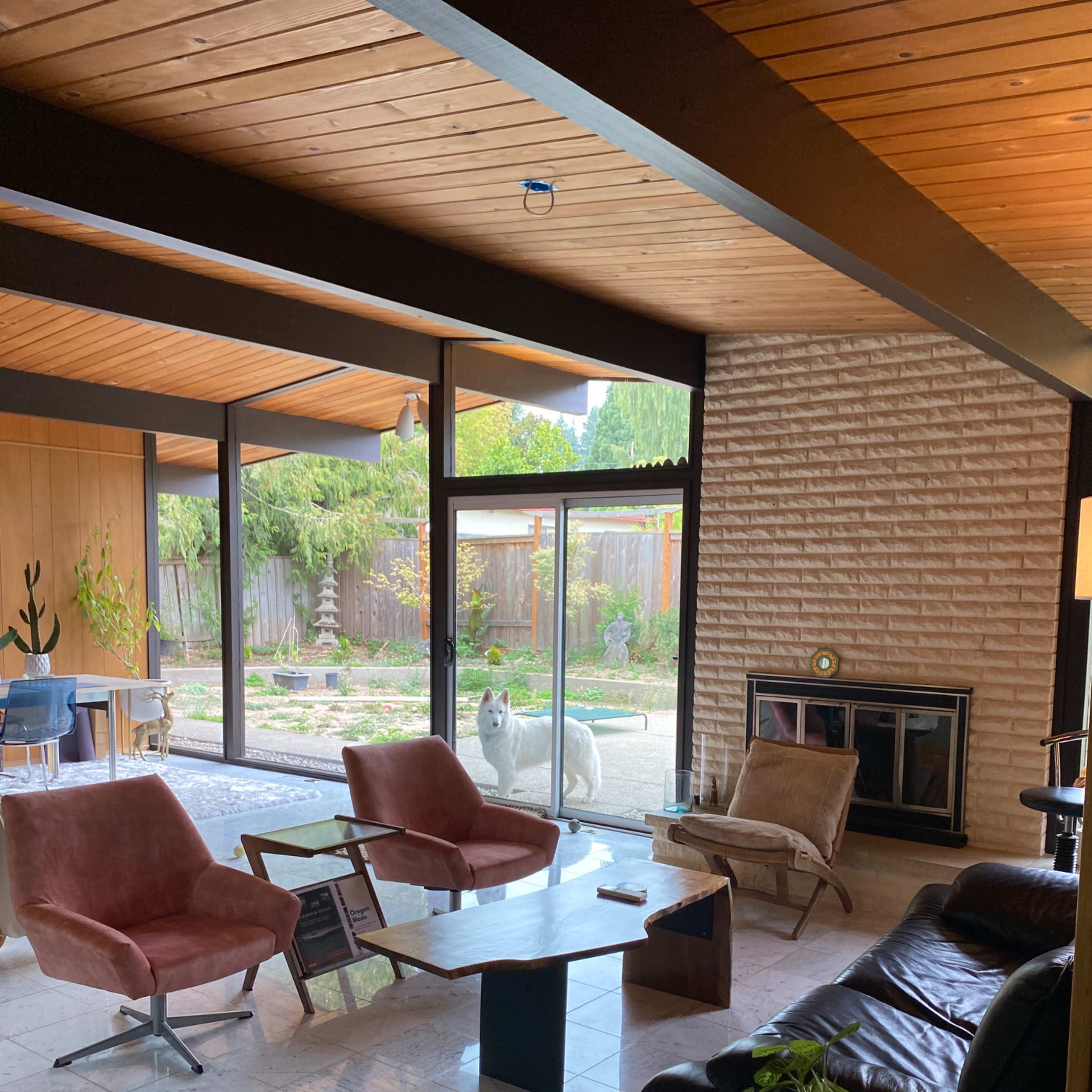 A mid-century modern living room with large windows, featuring a mix of seating options, a coffee table, and a view of a garden outside.