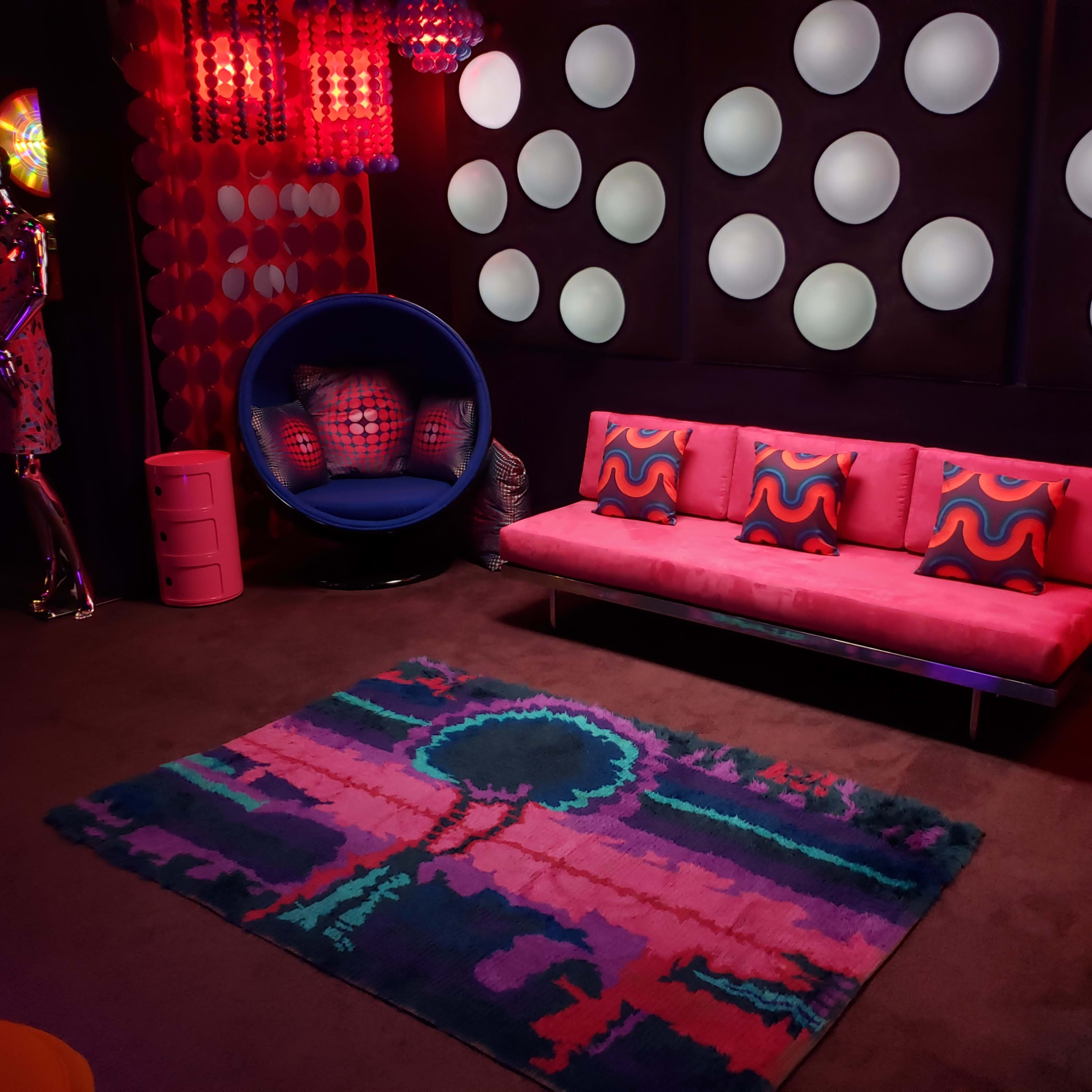 The room features a pink sofa with patterned cushions, a blue chair, colorful lighting, and a large rug with a tree design against a backdrop of circular wall decorations.