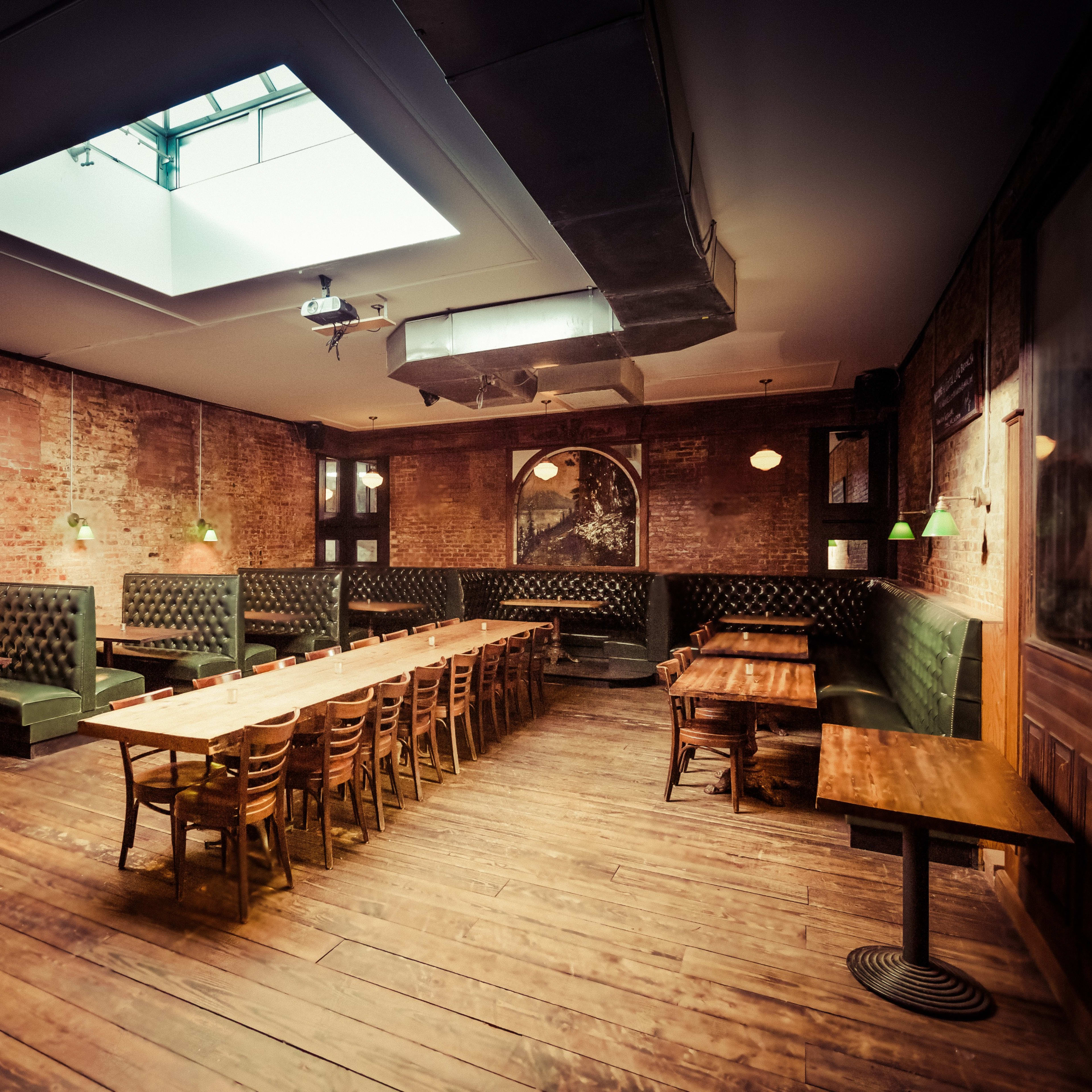 The interior of a restaurant features exposed brick walls, wooden floors, and a series of long wooden tables with green upholstered seating along the walls, illuminated by green pendant lights.