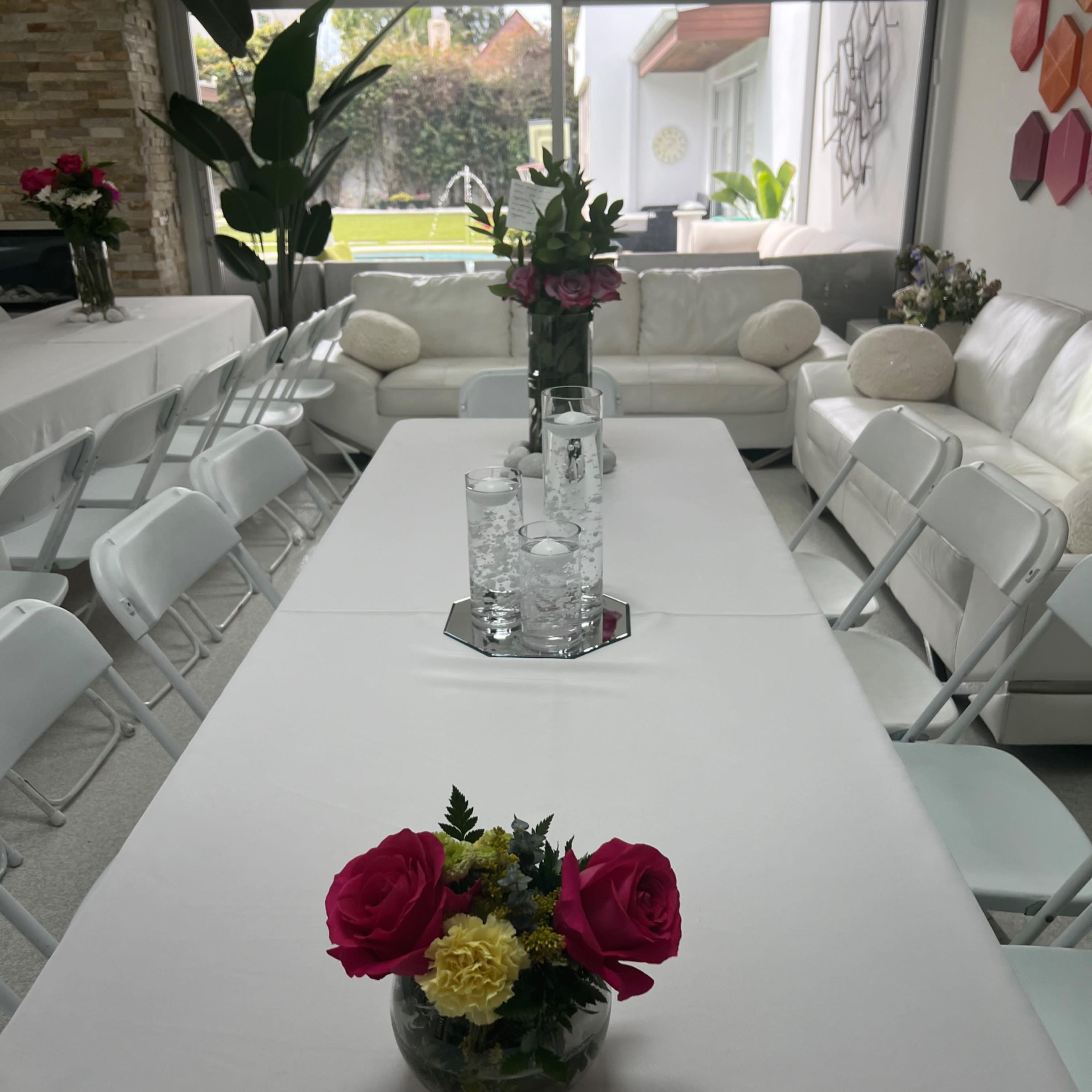 The scene shows a dining area set with white tables and chairs, adorned with floral centerpieces, and a view of a garden through large windows.