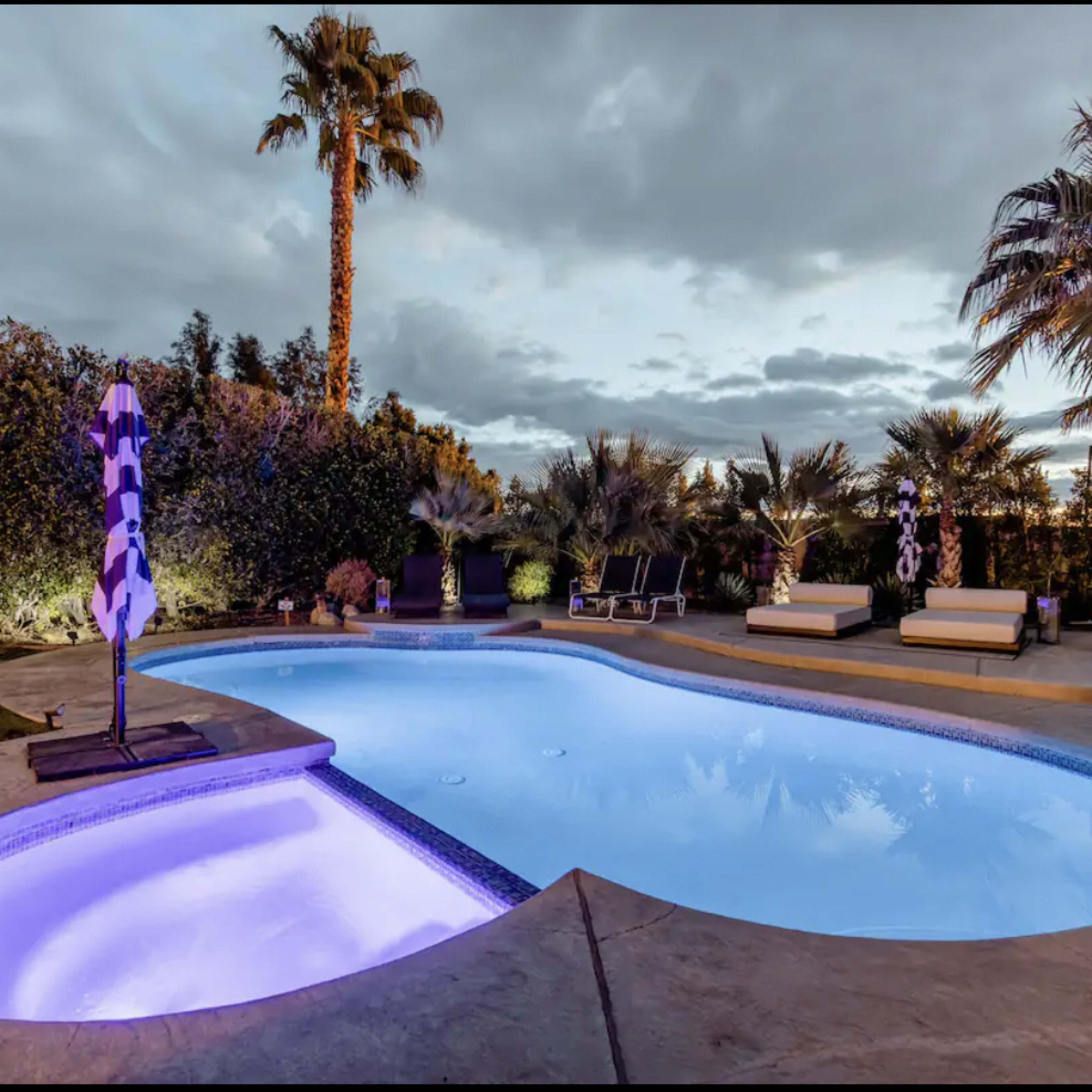 A swimming pool with illuminated water, surrounded by palm trees, lounge chairs, and a grassy area under a partly cloudy sky.