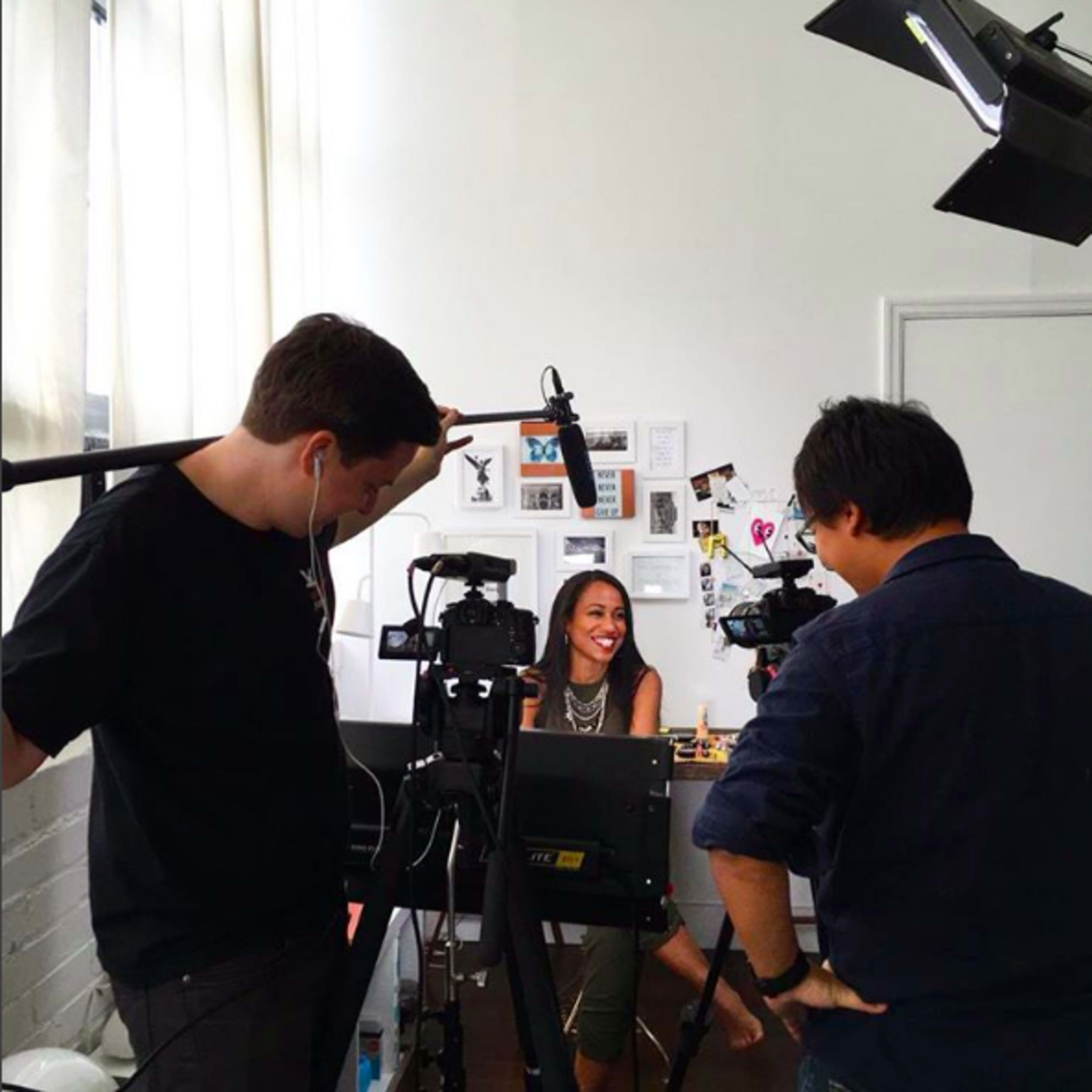 A woman is seated in front of a desk while being filmed by a crew with multiple cameras and microphones.