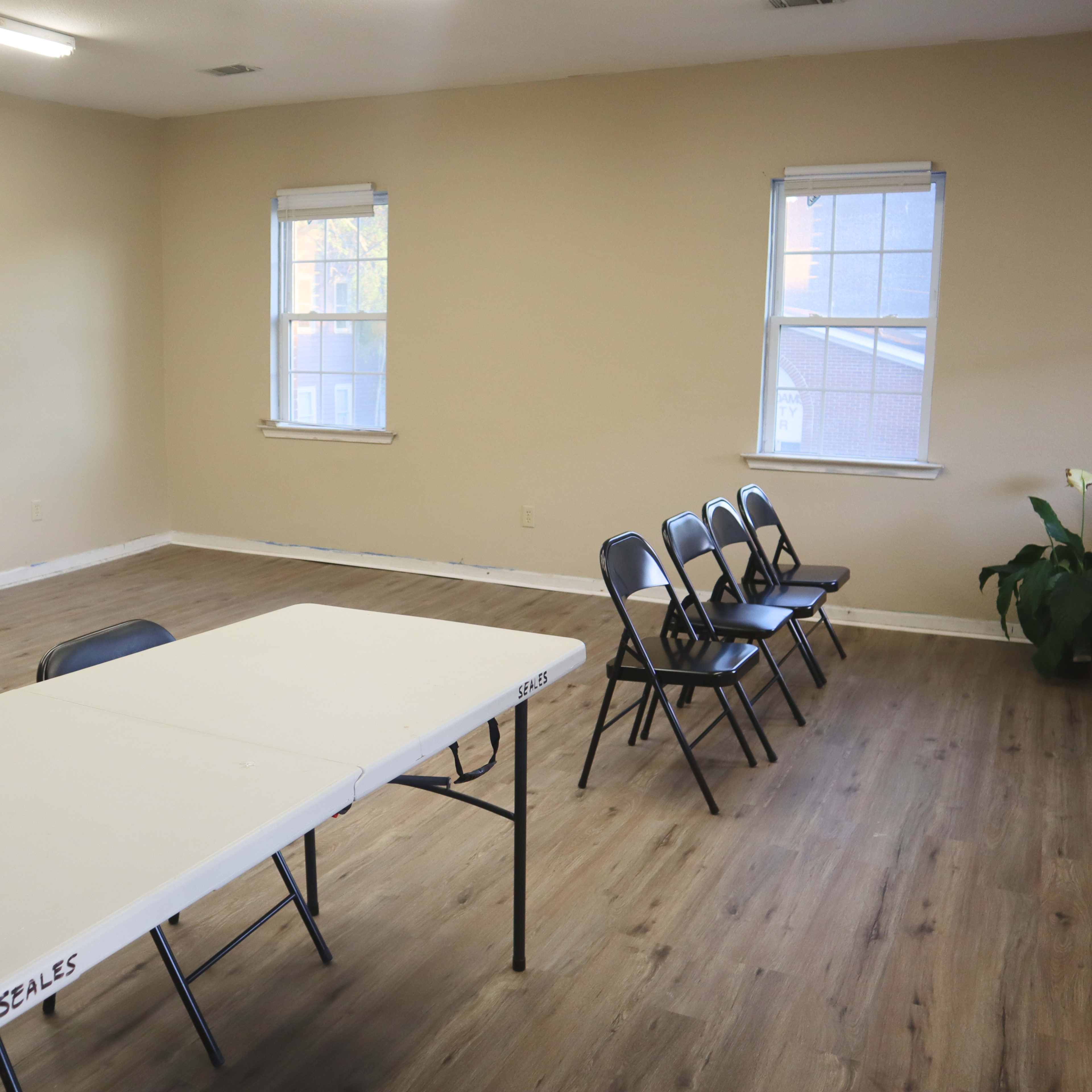 A sparsely furnished room with a long table and several chairs arranged in front of two windows.