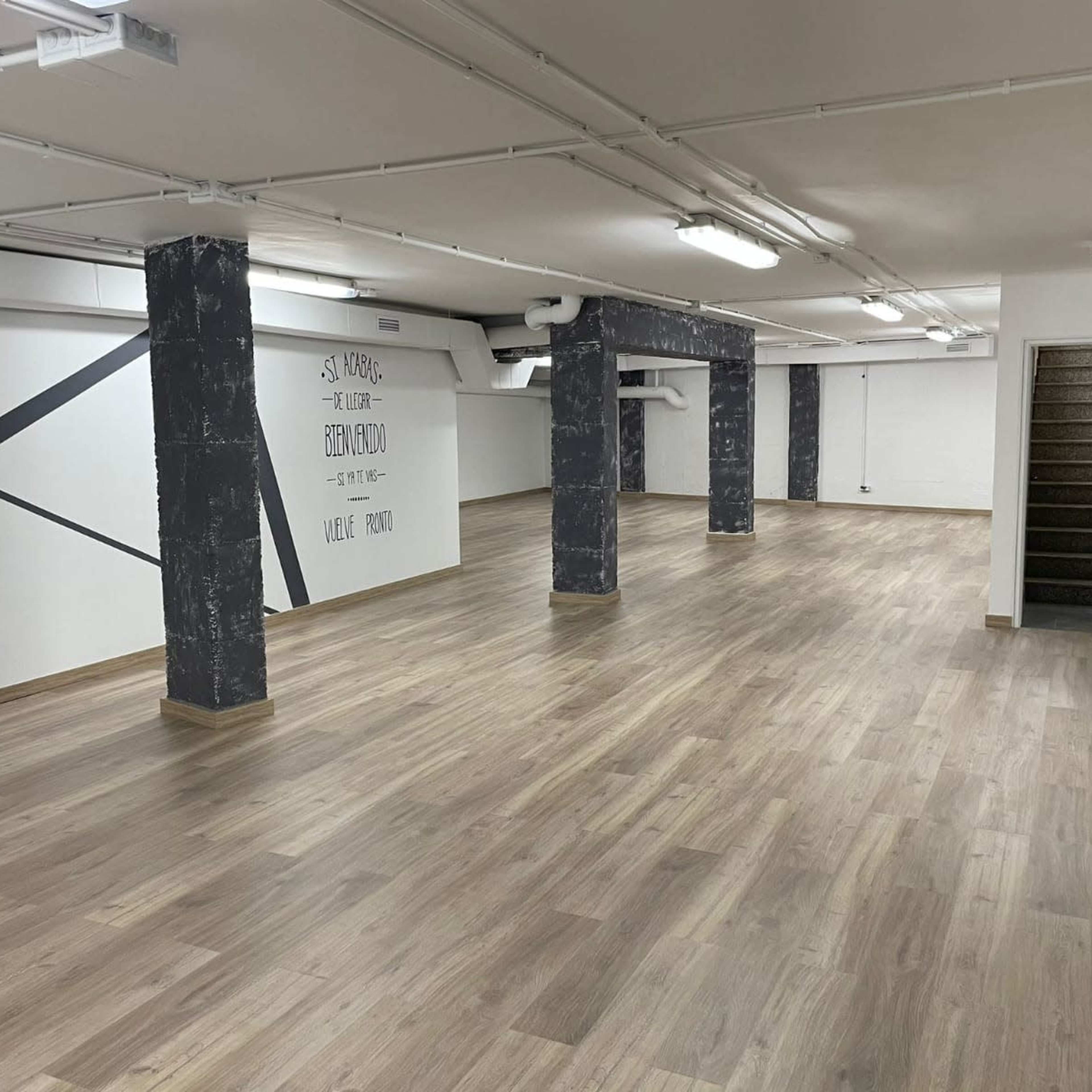 A spacious, empty indoor area with wooden floors, white walls featuring black geometric lines, and concrete columns, along with a set of stairs in the corner.