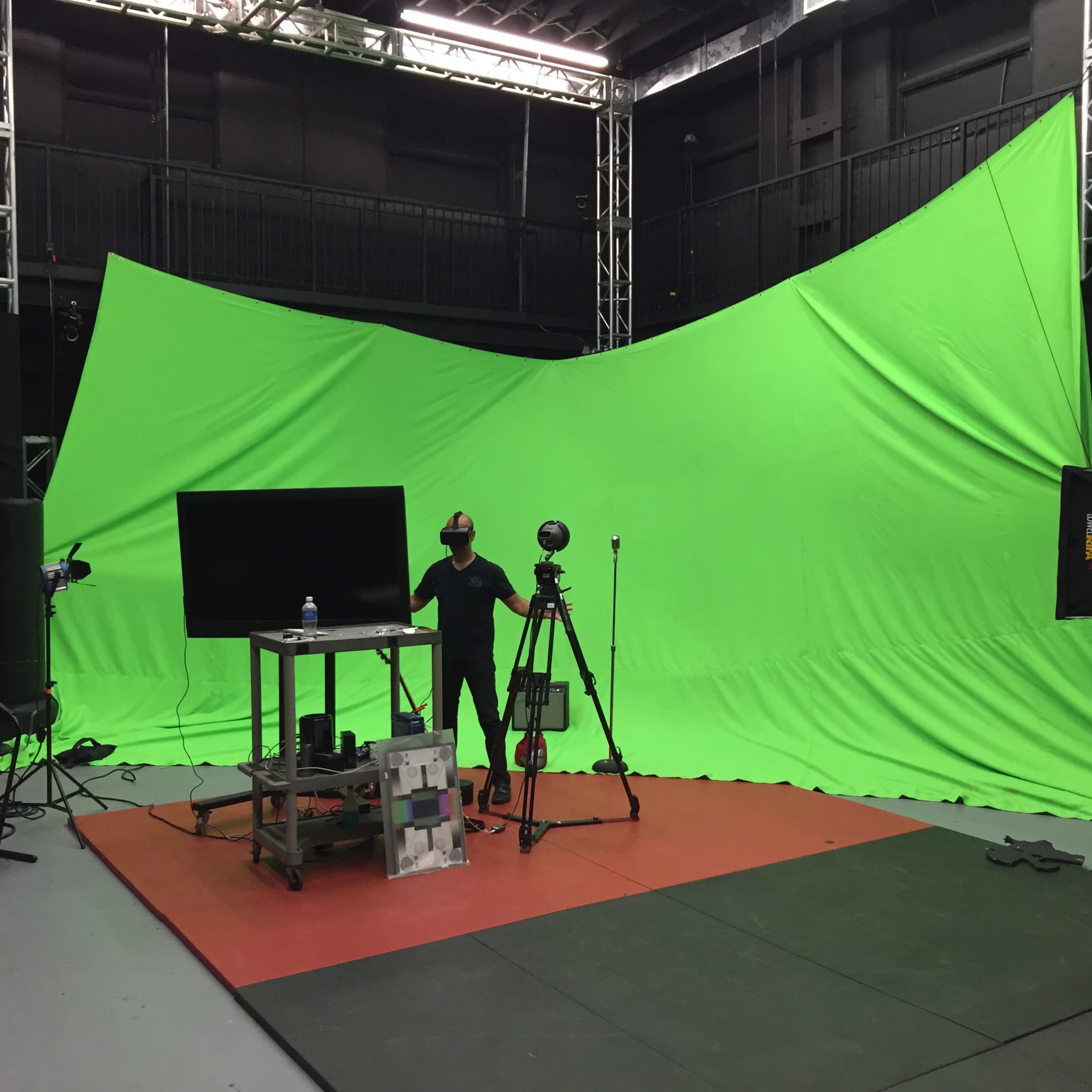 A green screen studio setup features a large green backdrop, a table with equipment, cameras, and lighting in an indoor space.