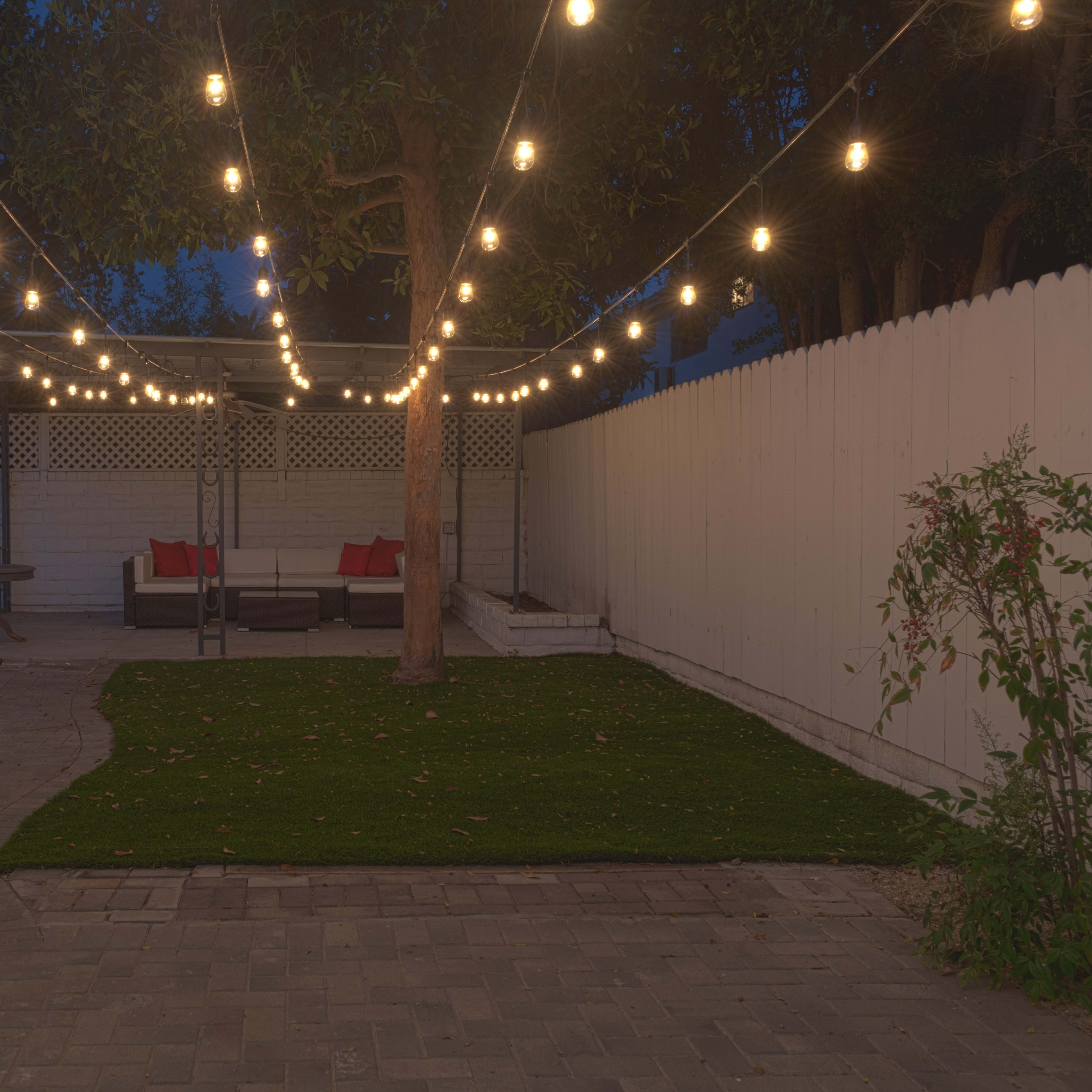 A backyard with string lights illuminating a seating area with red cushions, surrounded by a lawn and a white fence.