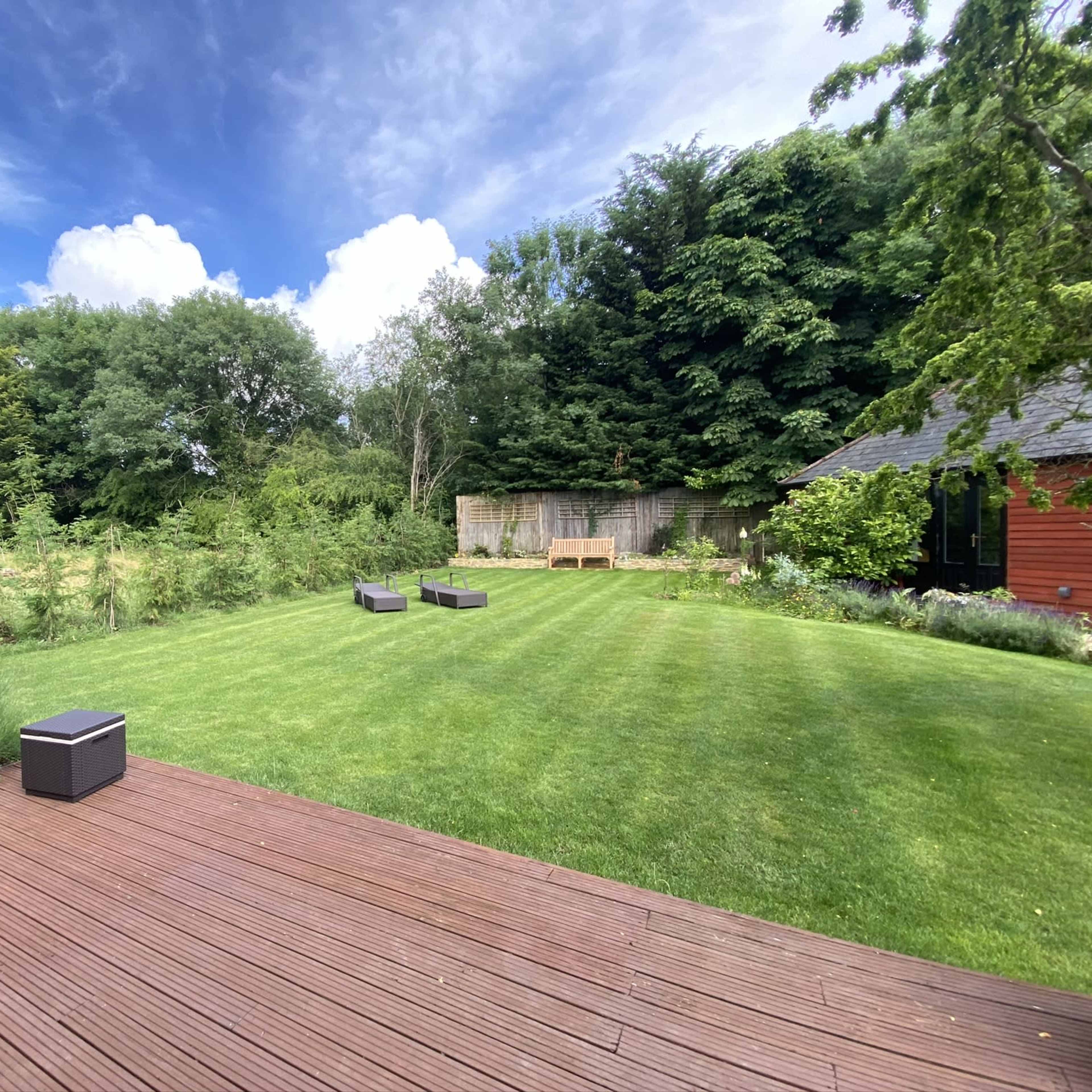A well-maintained lawn features two seating areas and is surrounded by trees and shrubs under a partly cloudy sky.