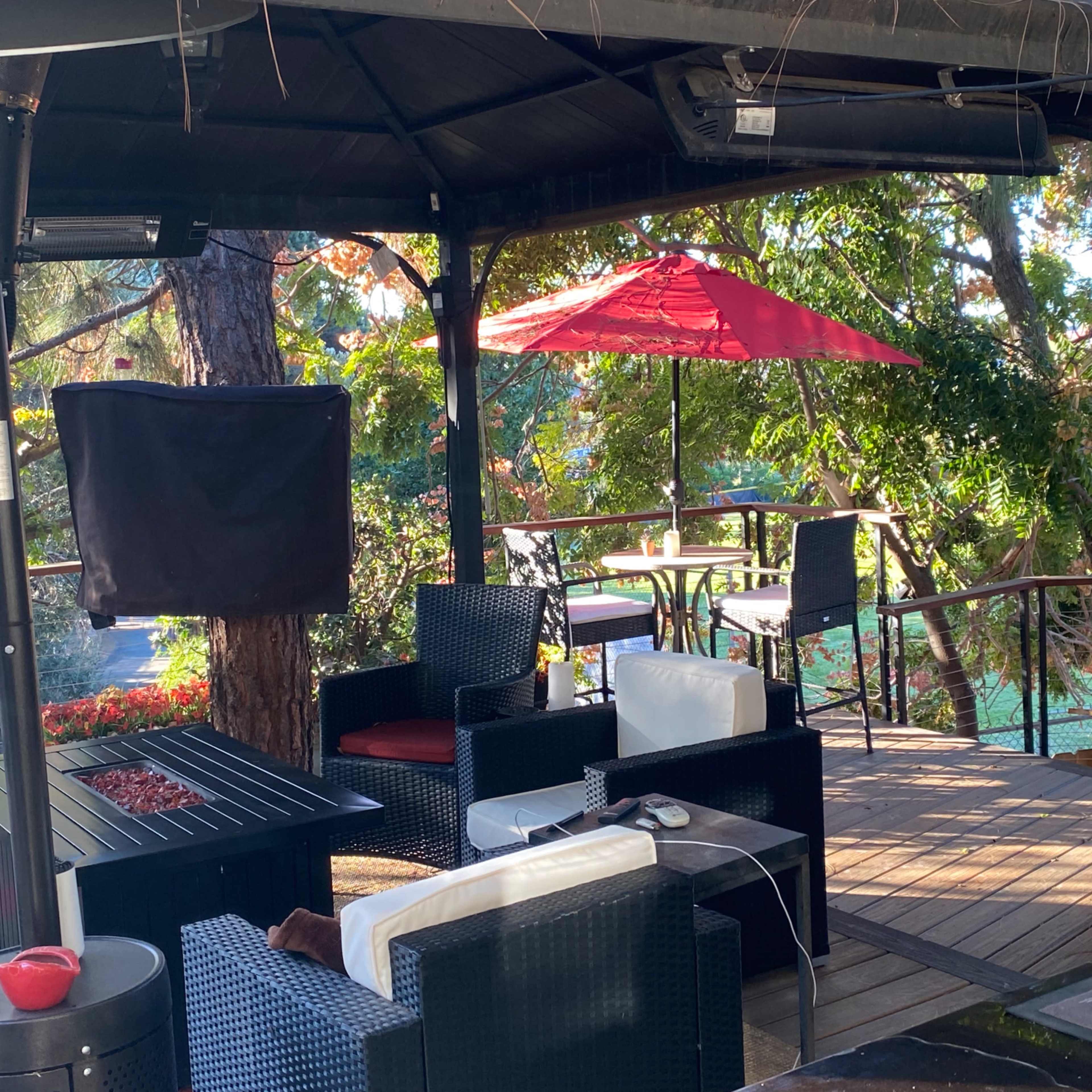 A covered outdoor seating area with black wicker furniture and a red umbrella on a wooden deck surrounded by trees.