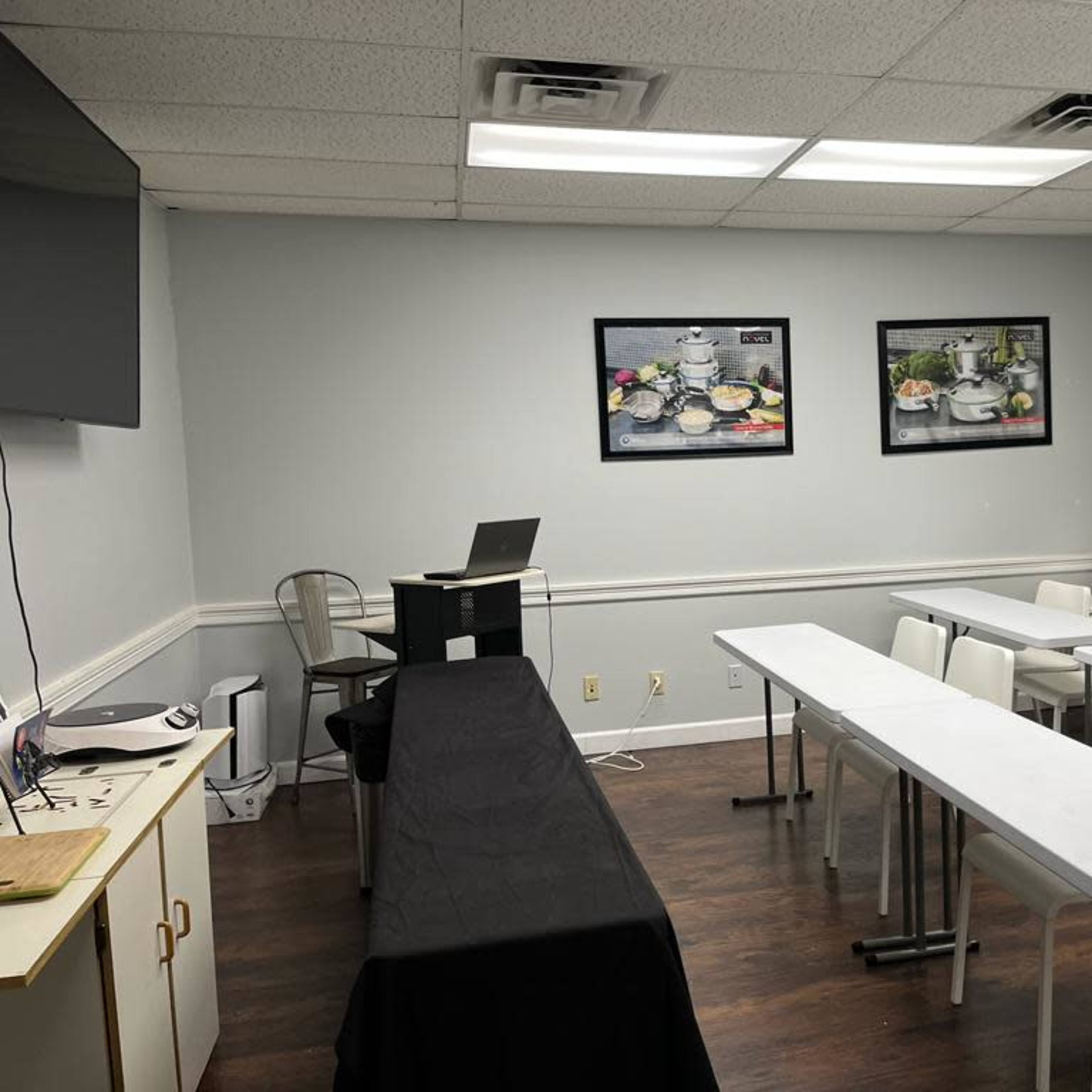 A small, well-lit meeting room features a black table at the front, rows of white tables and chairs, and two framed pictures on the walls.