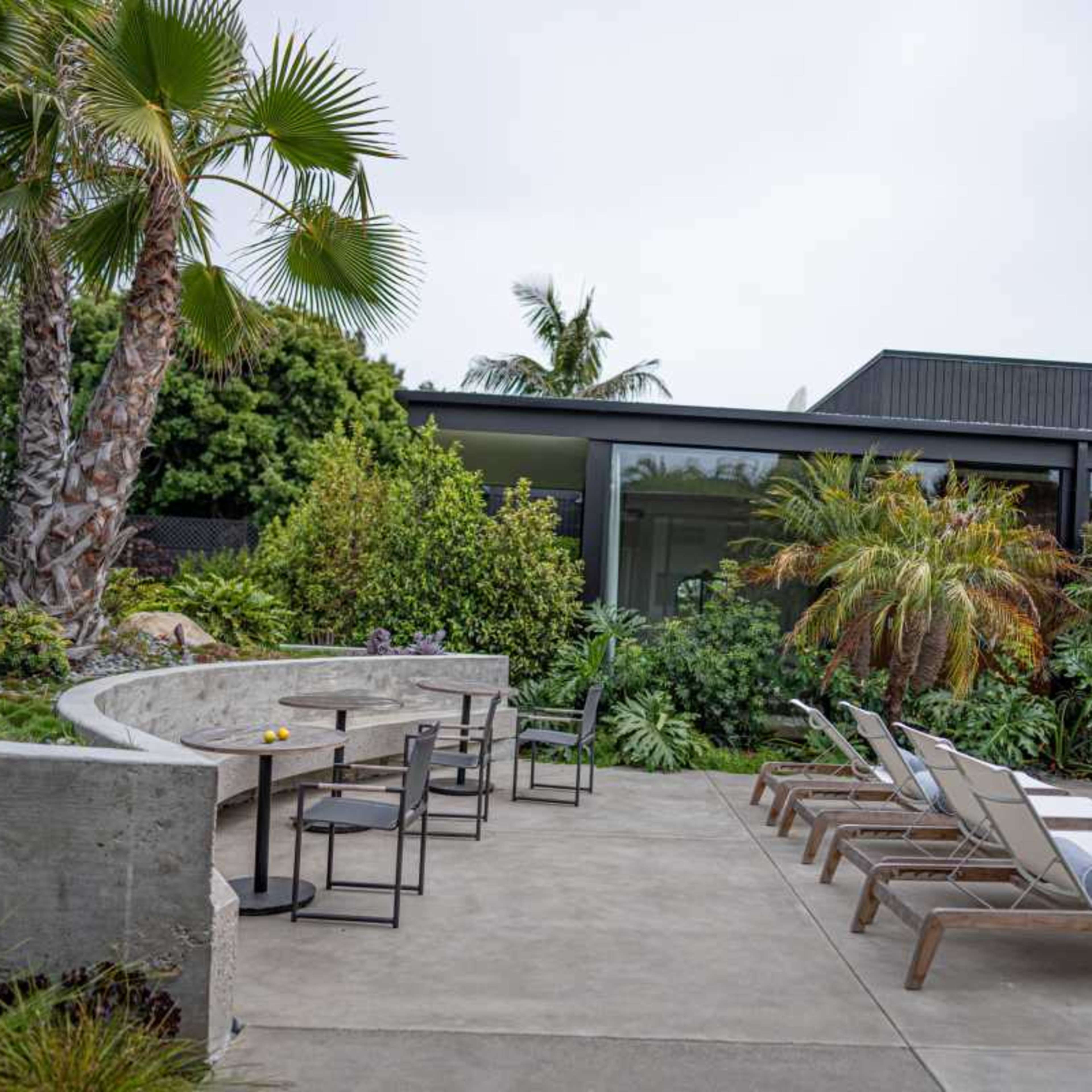A poolside area features lounge chairs, a concrete seating arrangement, and lush tropical plants surrounded by a modern building.