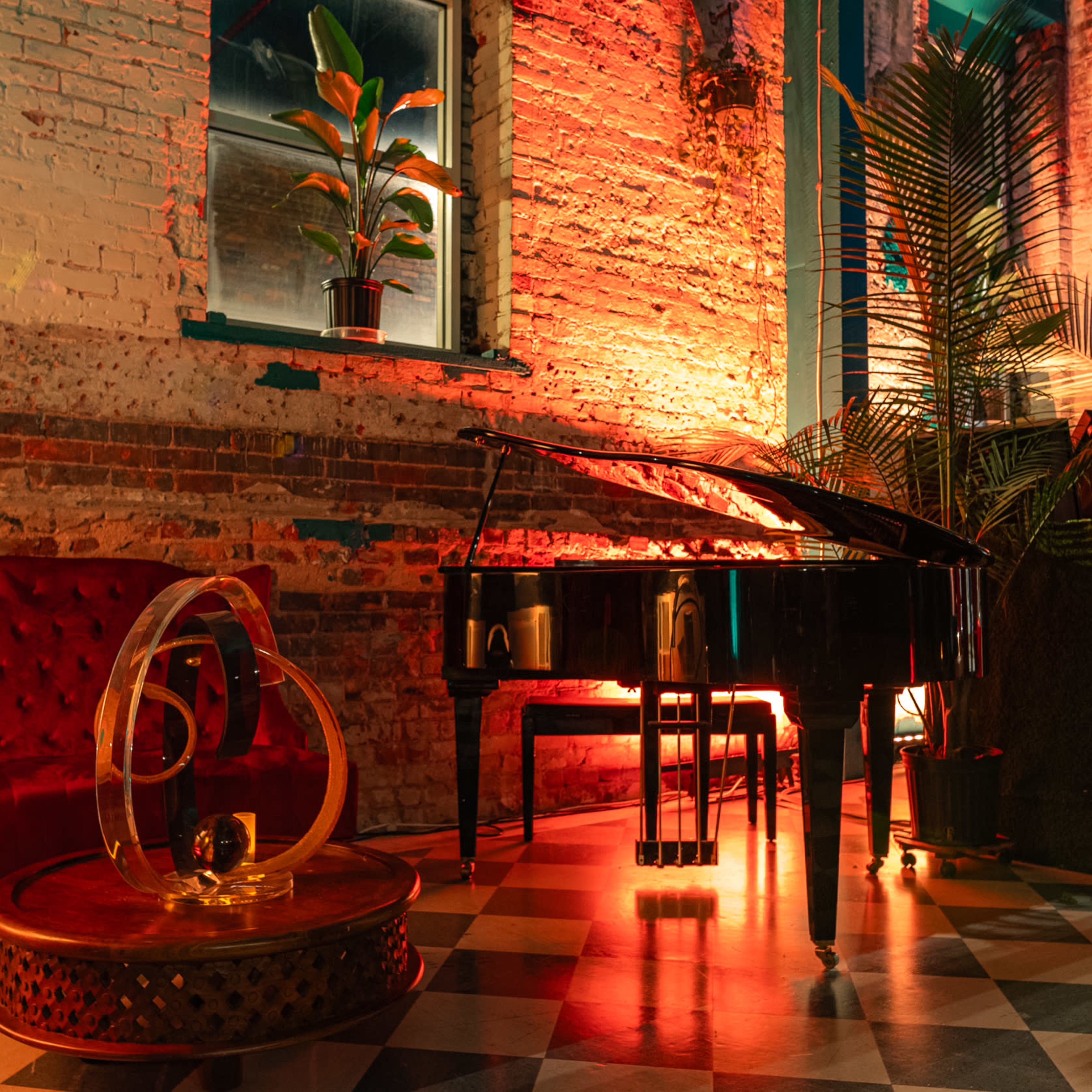 A grand piano is positioned next to a red velvet sofa and a round wooden table, surrounded by plants and illuminated by warm lighting in a brick-walled setting.