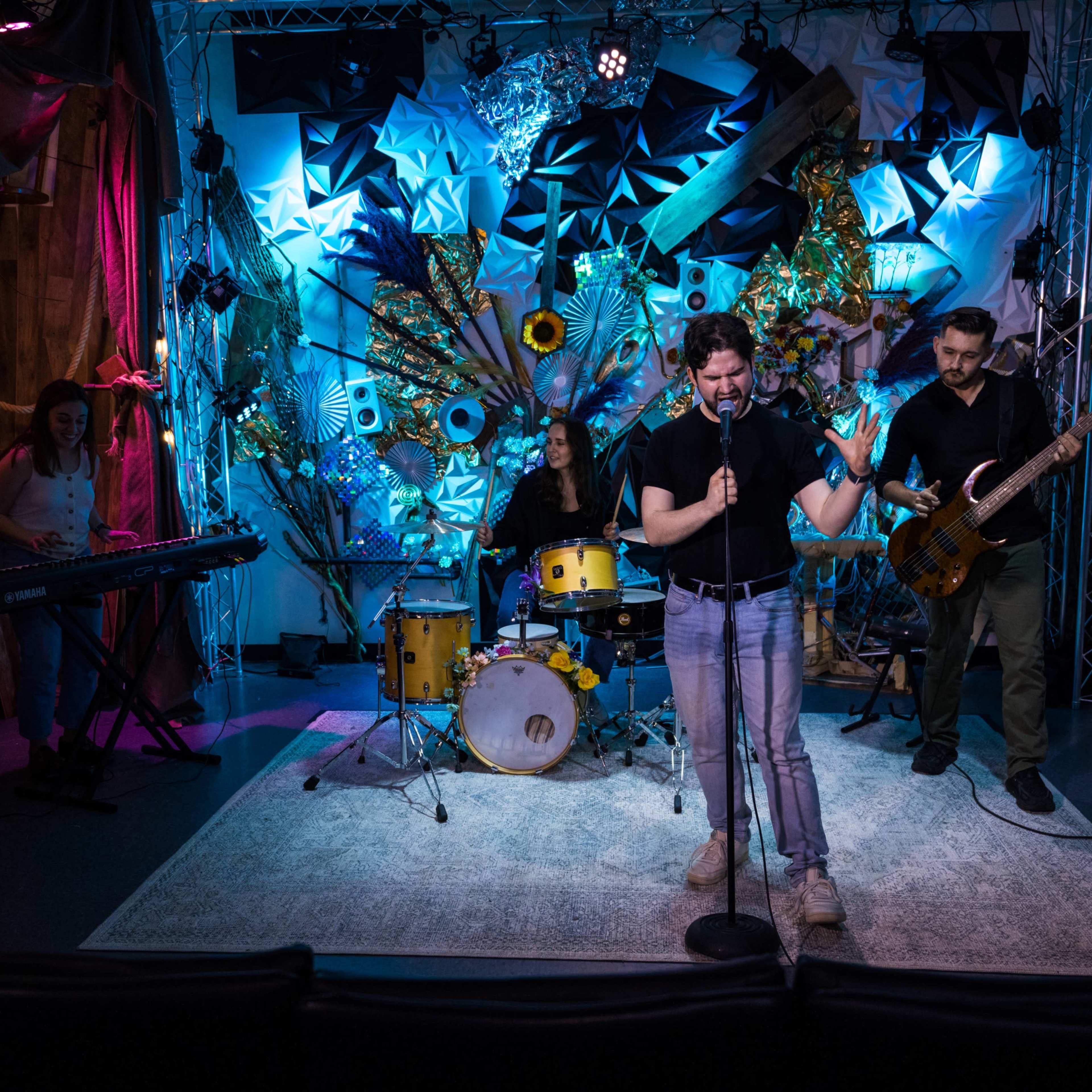 A band performs on stage in a colorful, creatively decorated room with various textured backdrops and lighting.