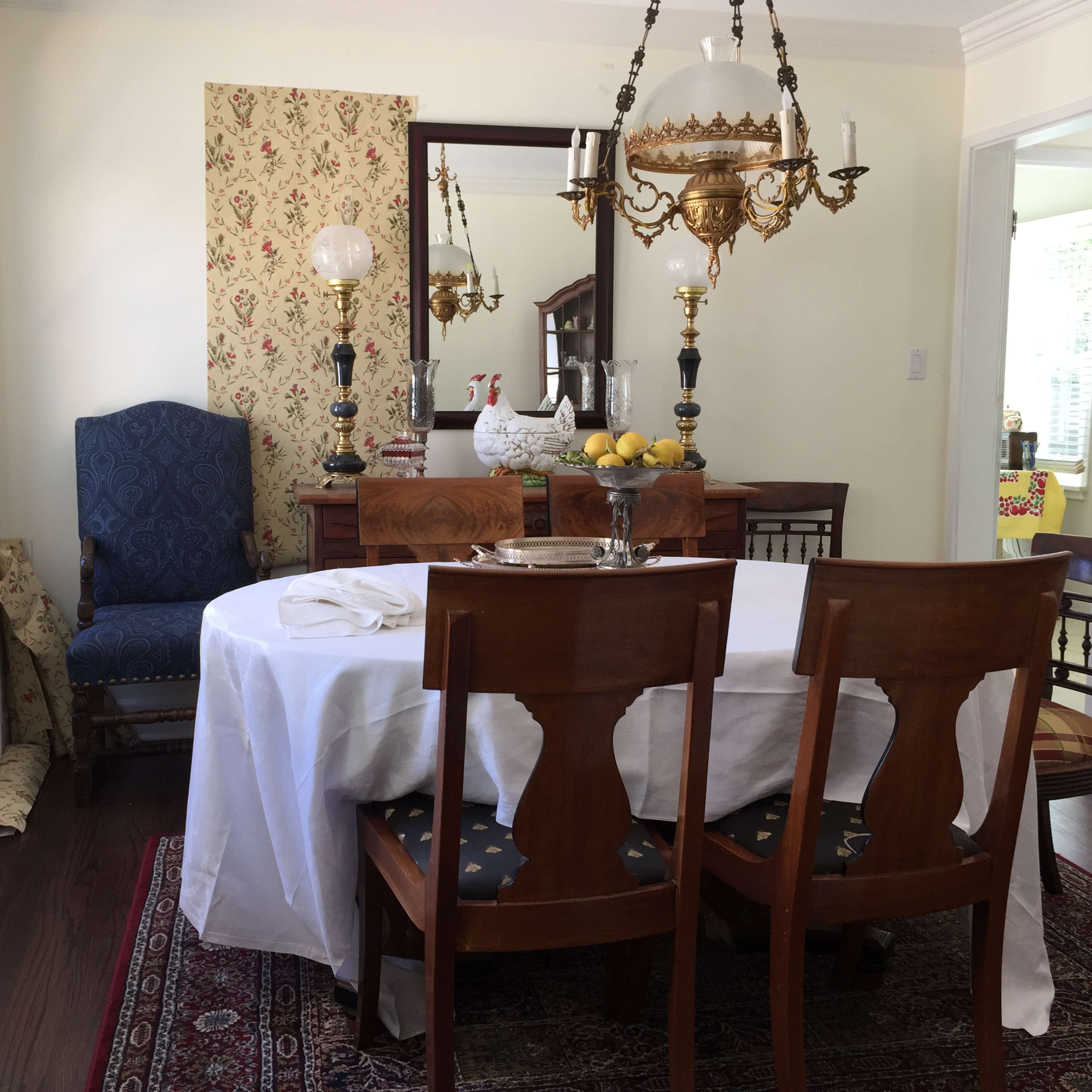 A dining room features a round table covered with a white tablecloth, surrounded by wooden chairs, and a chandelier hangs above.