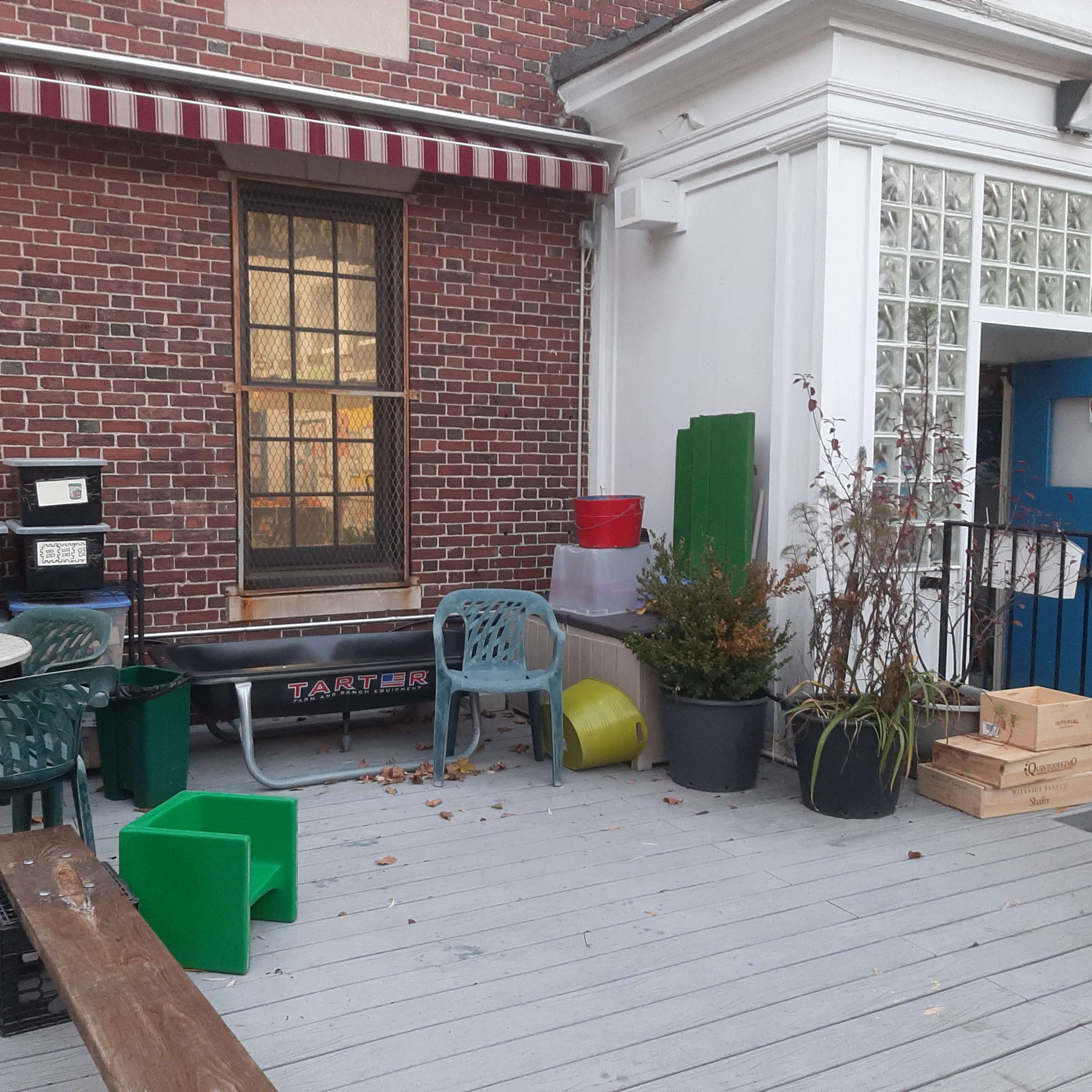A small outdoor area with a wooden deck, various chairs and tables, plant pots, and storage containers against a brick wall.