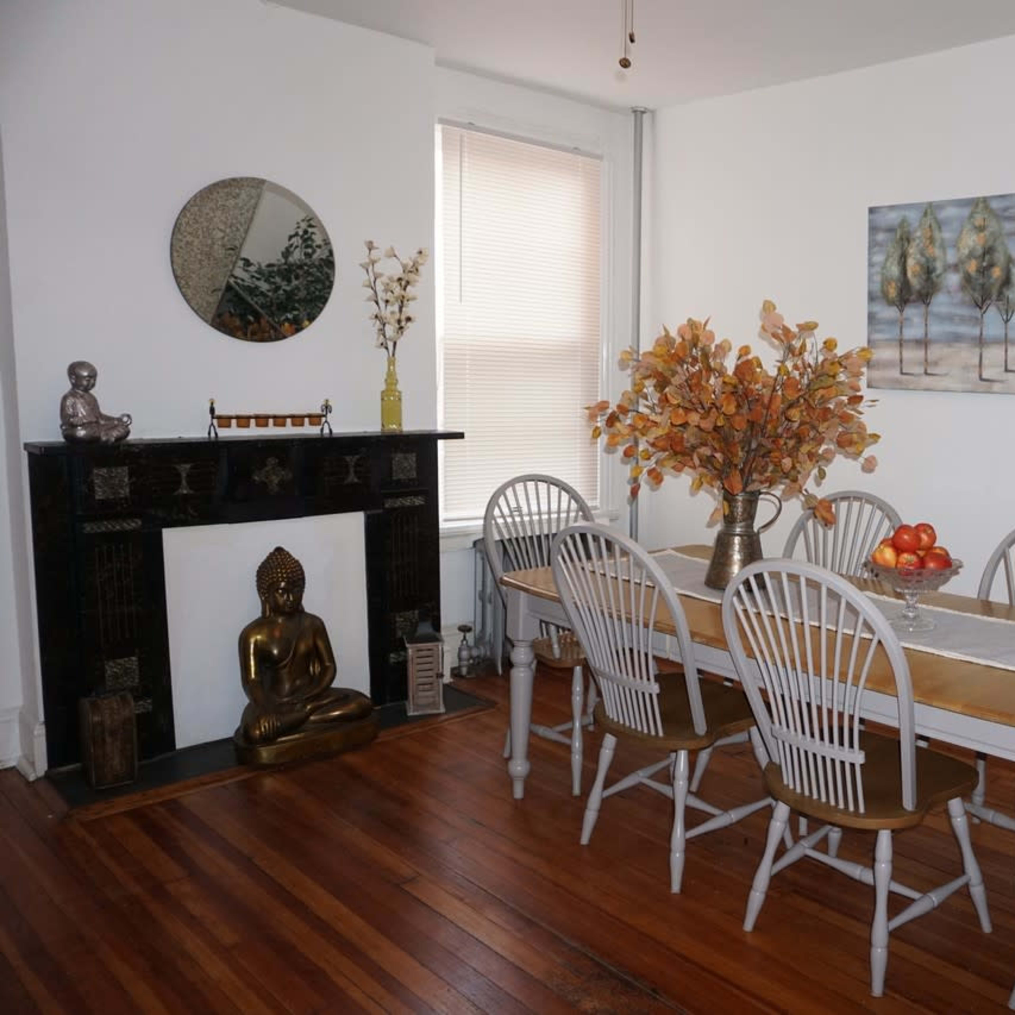 A dining room features a long table surrounded by wooden chairs, with a decorative centerpiece of autumn foliage and a statue of a seated figure on a mantel.