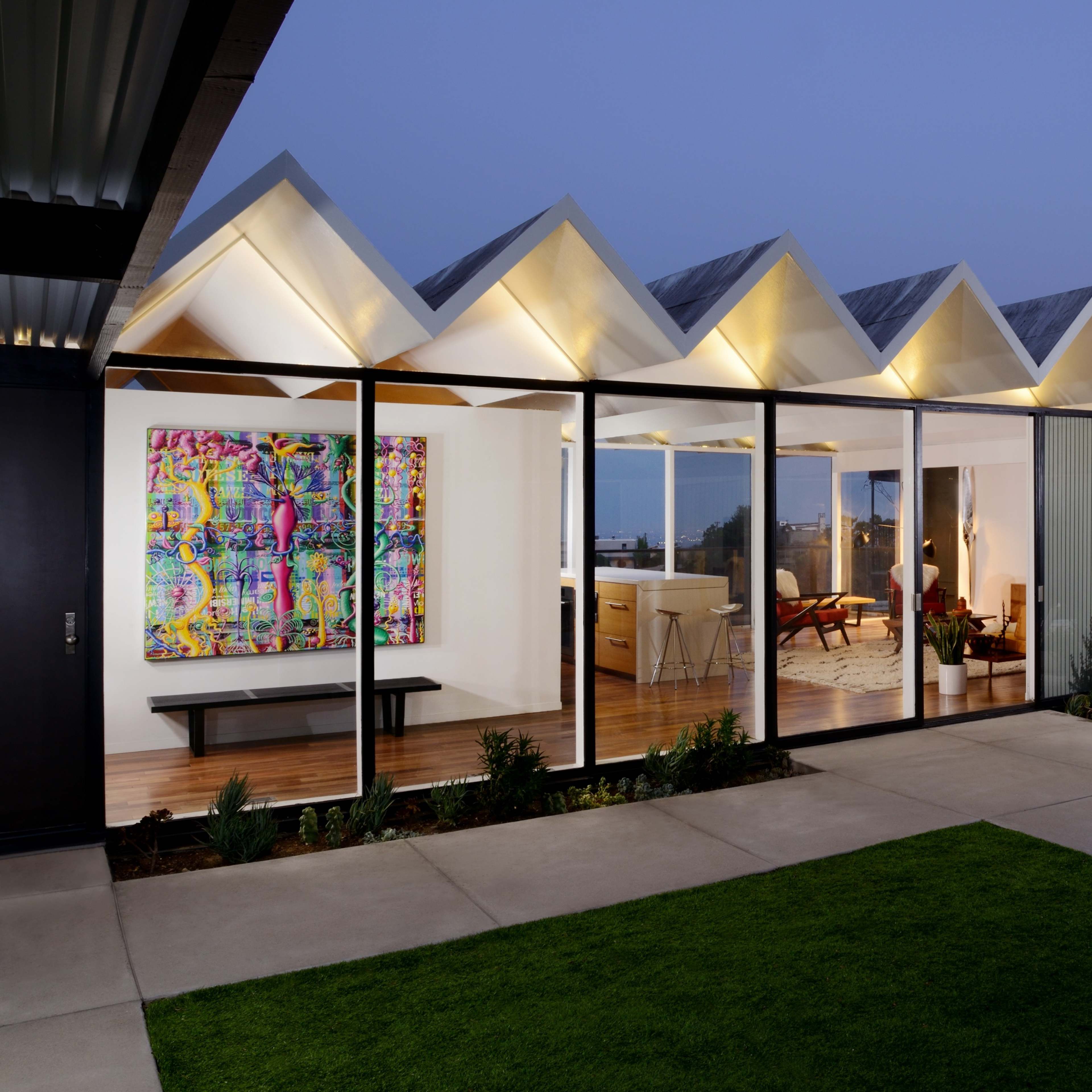 A modern house with a distinctive zigzag roof features large glass walls, showcasing an open interior and a backyard with a well-maintained lawn.