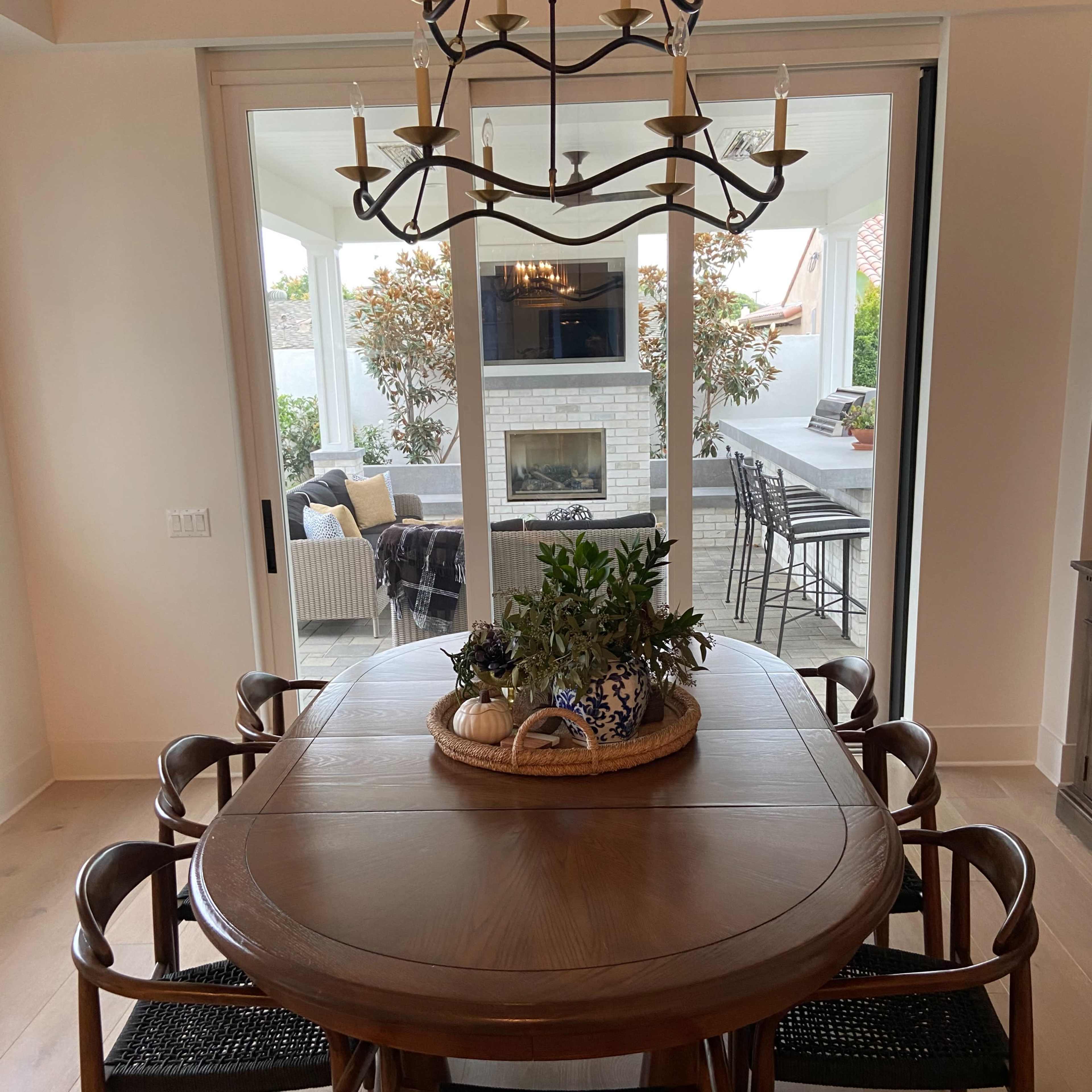 A wooden dining table with a central plant arrangement is surrounded by six chairs, and large glass doors lead to a patio area with outdoor seating and a fireplace.