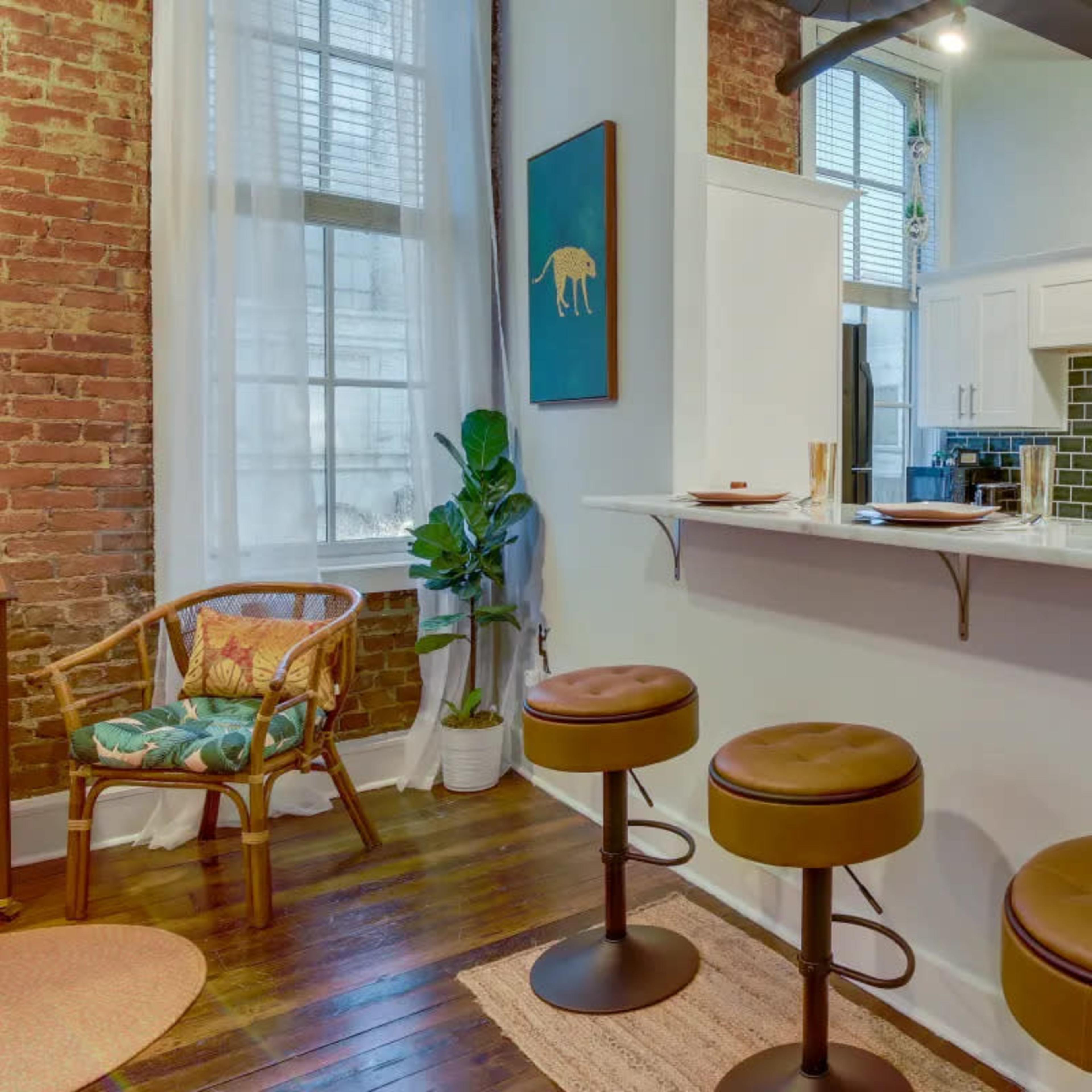 A modern kitchen area with a bar counter, three stools, and exposed brick walls, complemented by a small dining space with a chair and plant.