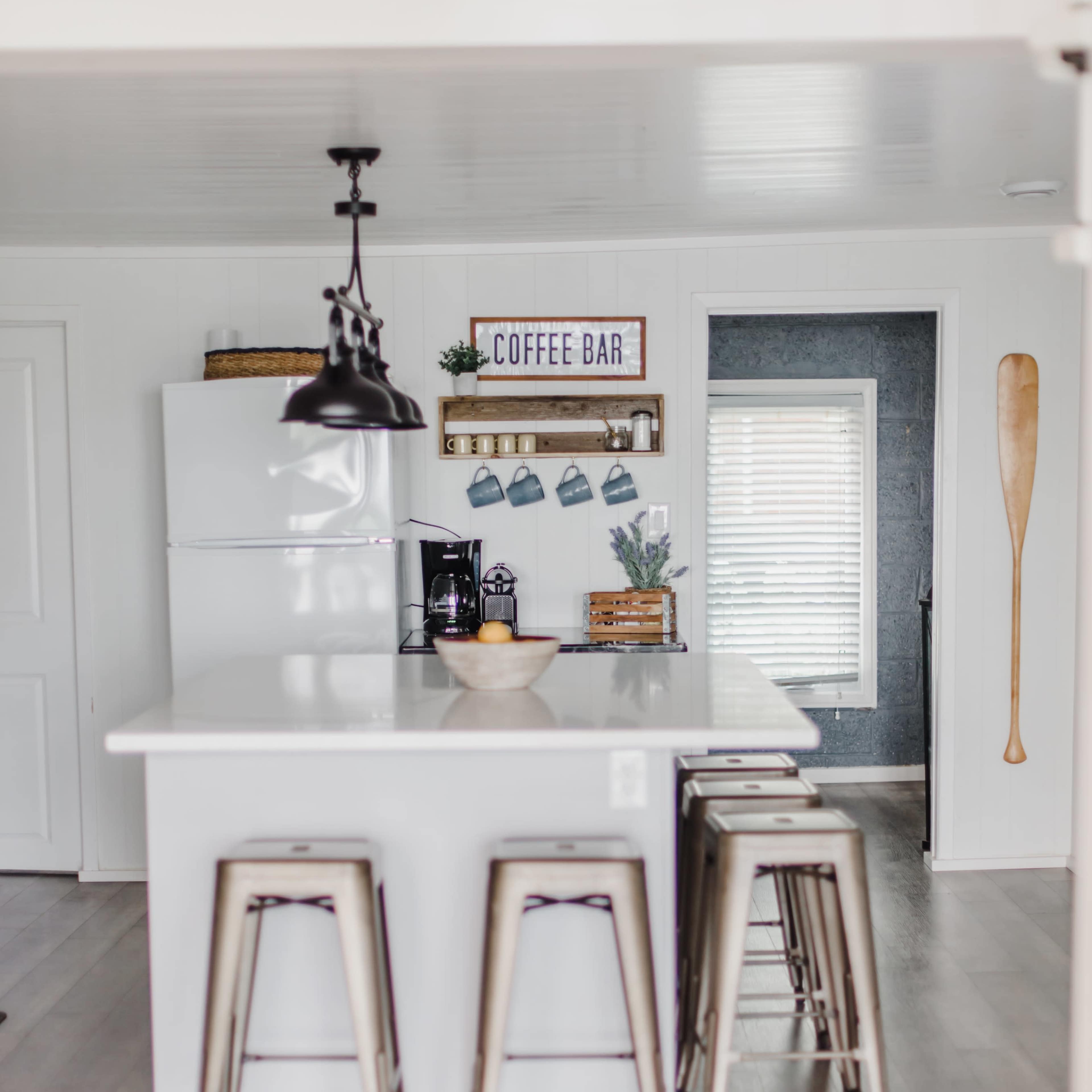 A modern kitchen features a central island with metal stools, a coffee bar against the wall, and stainless steel appliances.