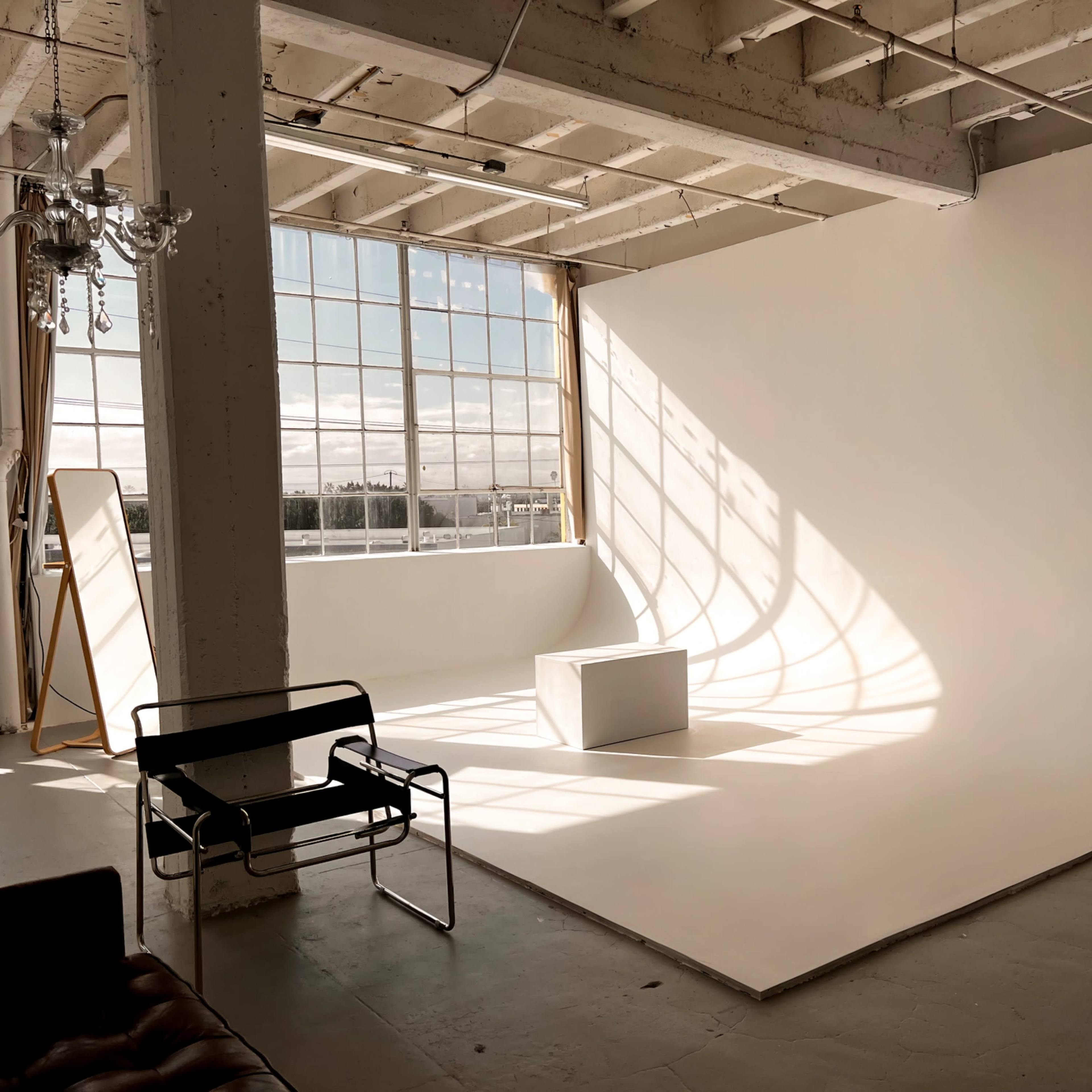 A spacious photography studio with a large white backdrop, natural light streaming through tall windows, and minimal furniture including a chair and a black metal chair.