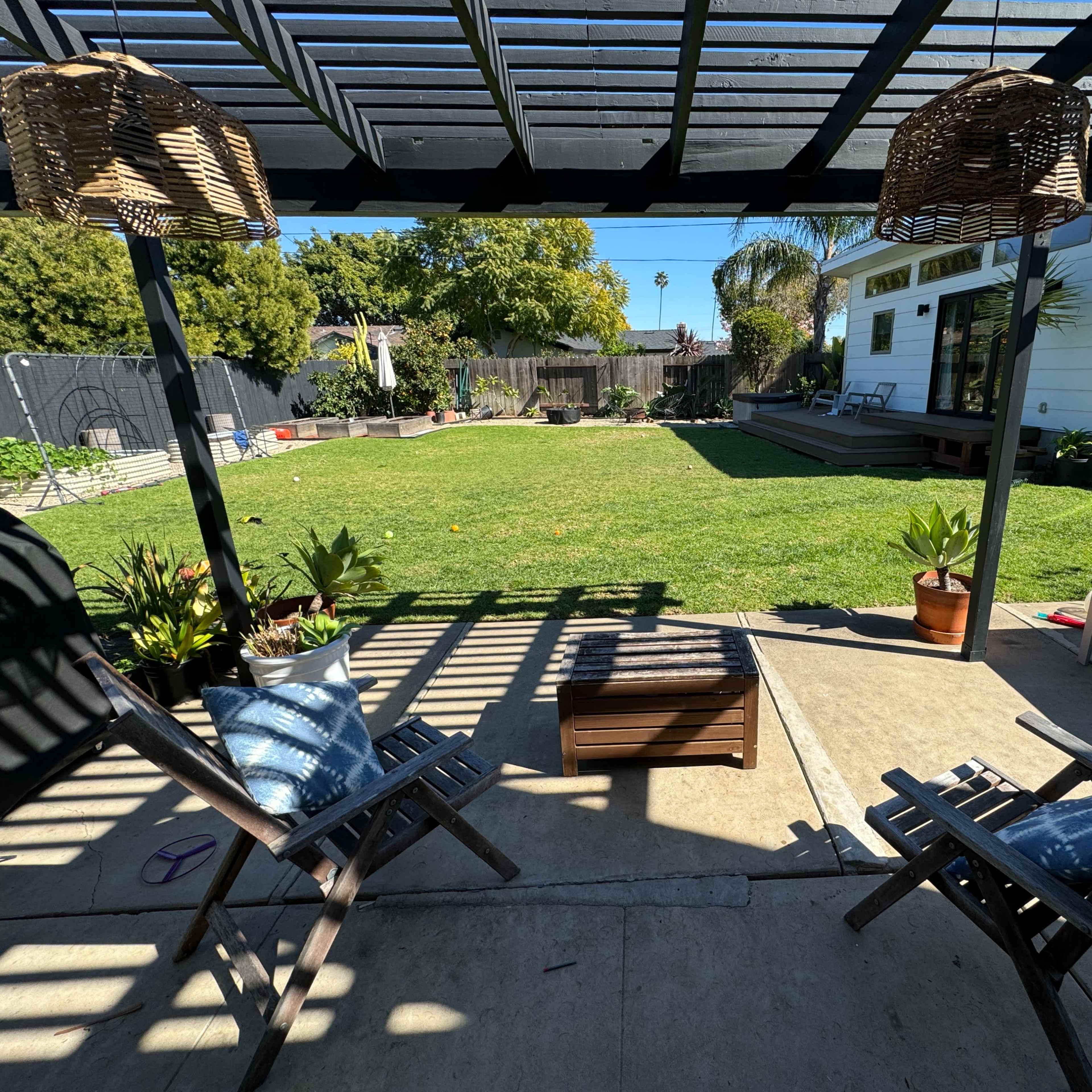 A backyard patio area with two wooden chairs, a small coffee table, and a well-maintained lawn surrounded by various plants and a house in the background.