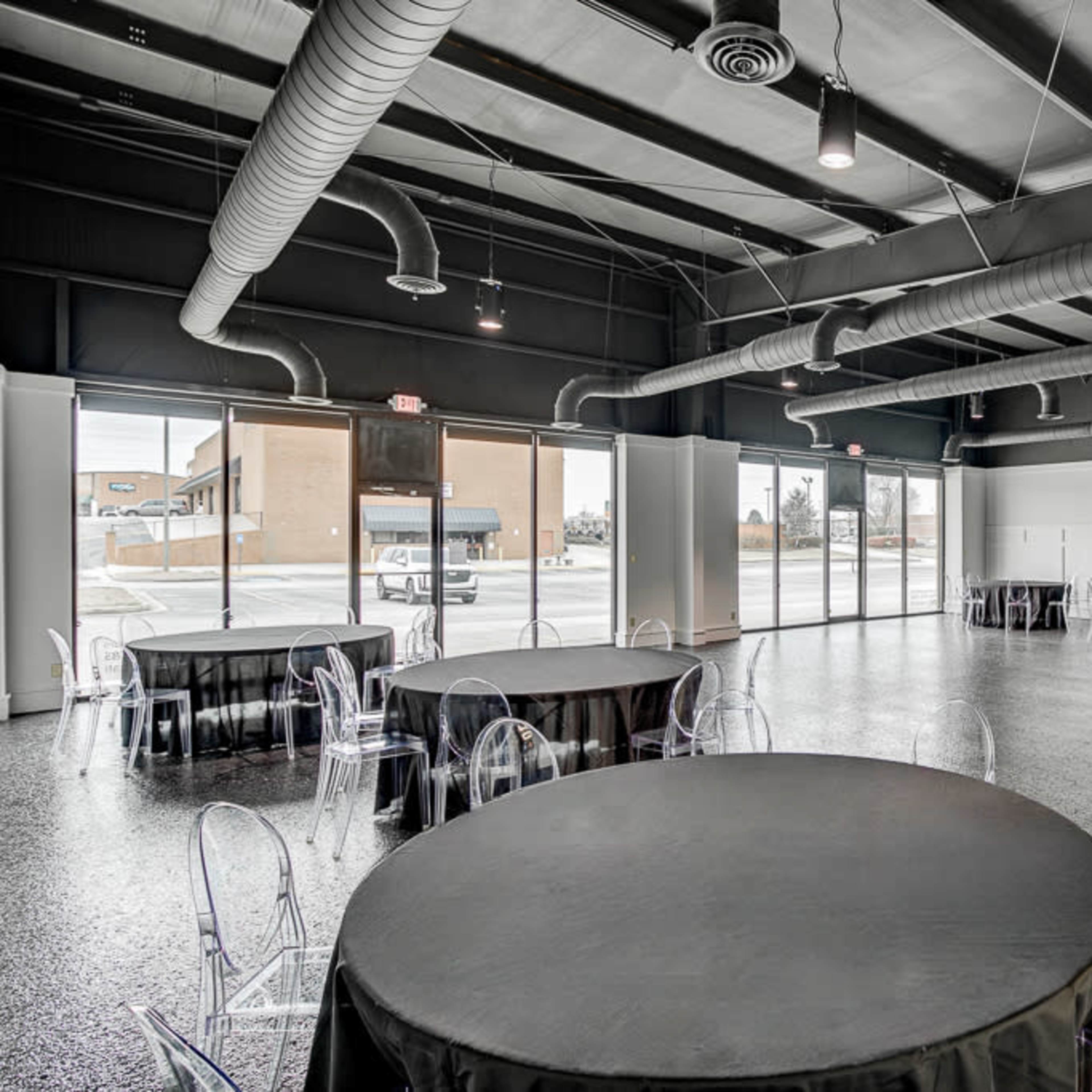 The interior of an event space features several round tables with black tablecloths and transparent chairs, illuminated by natural light from large windows.