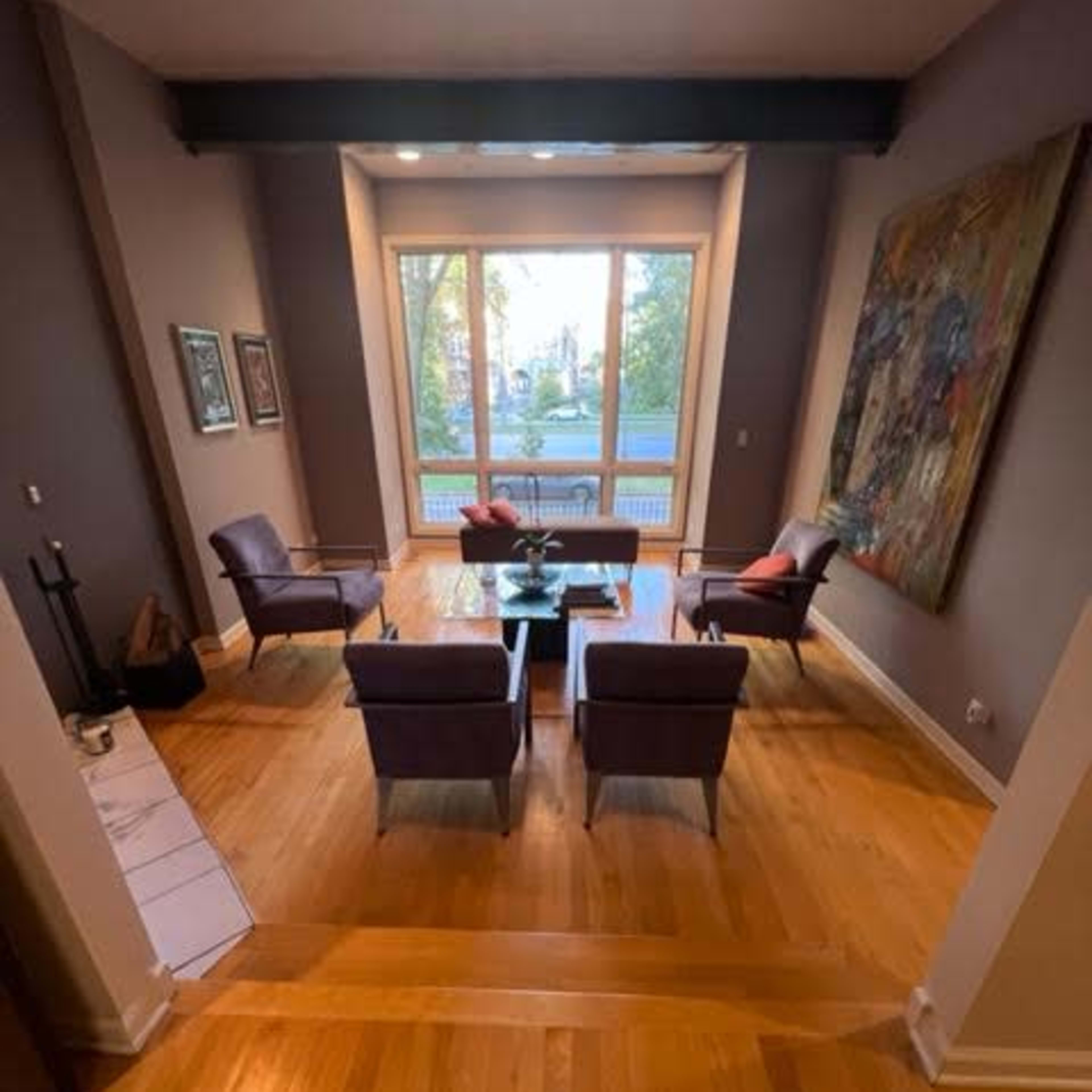 A spacious living area with hardwood floors, a central coffee table, and four chairs arranged around it, all set in front of large windows that provide natural light.