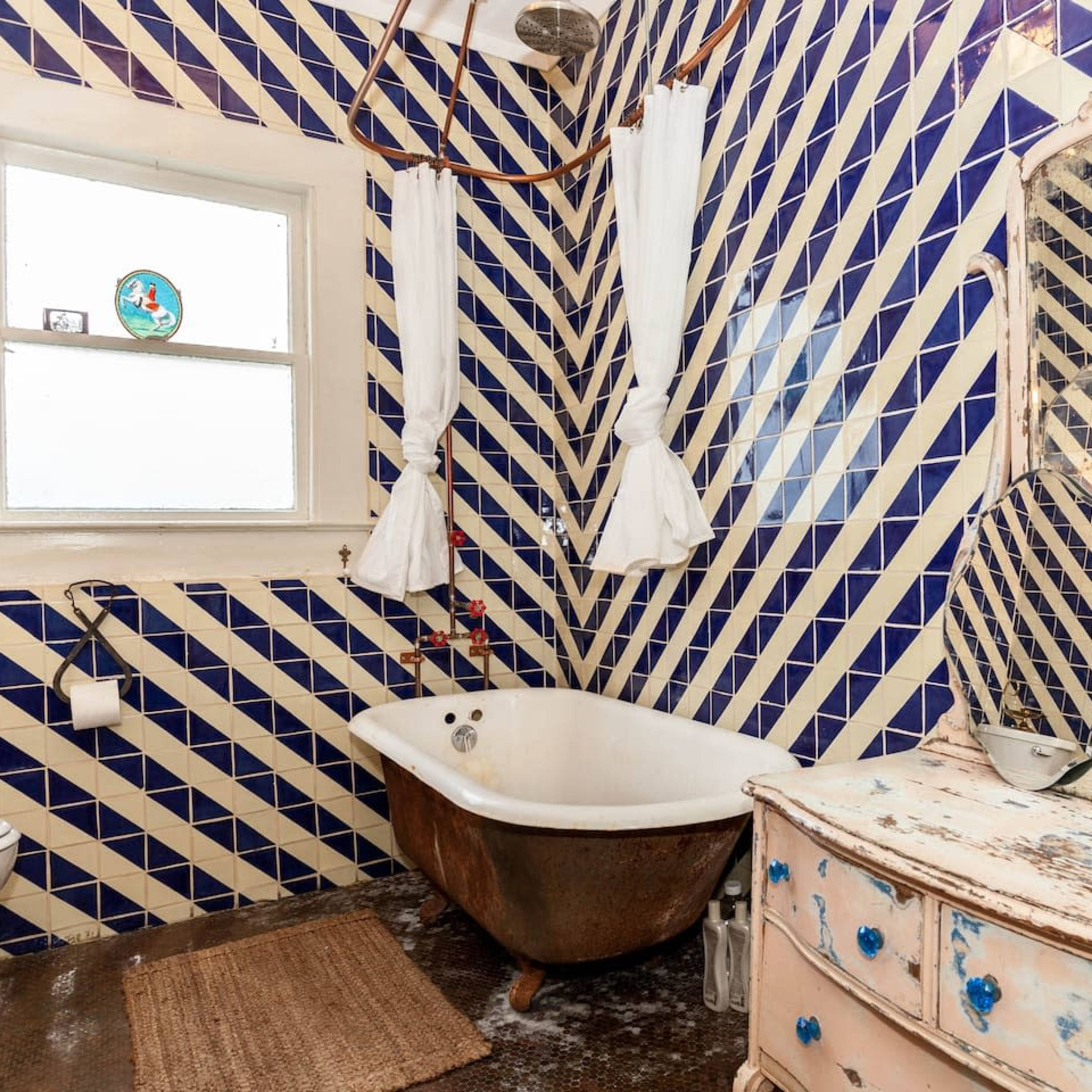 The bathroom features a clawfoot bathtub, a vintage mirror, and walls adorned with blue and white geometric tiles.
