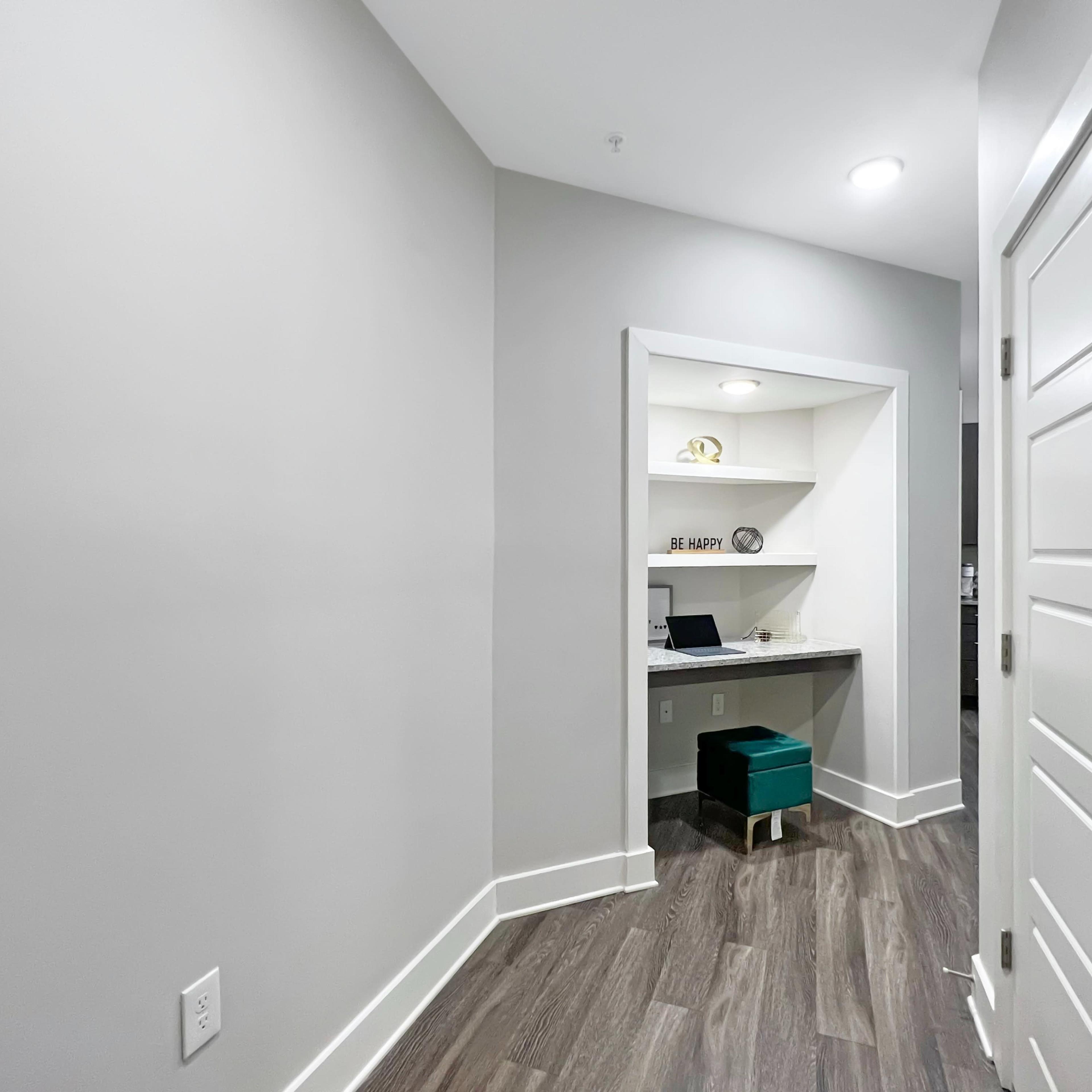 A small hallway leading to a nook with a desk and shelves, featuring a laptop and a decorative sign.