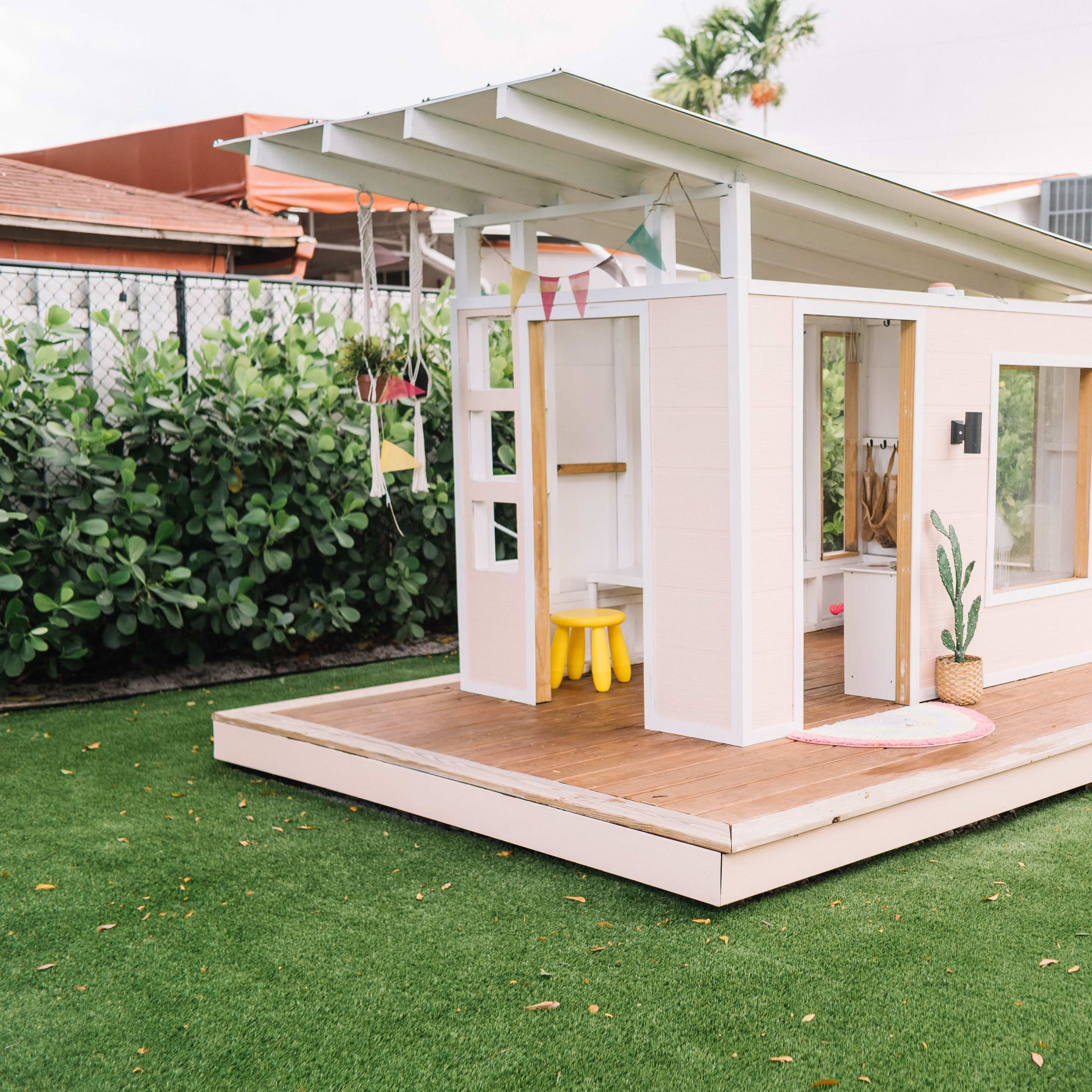 A small wooden playhouse with a slanted roof and a porch is situated on a grassy lawn, surrounded by greenery and pink flamingo lawn ornaments.