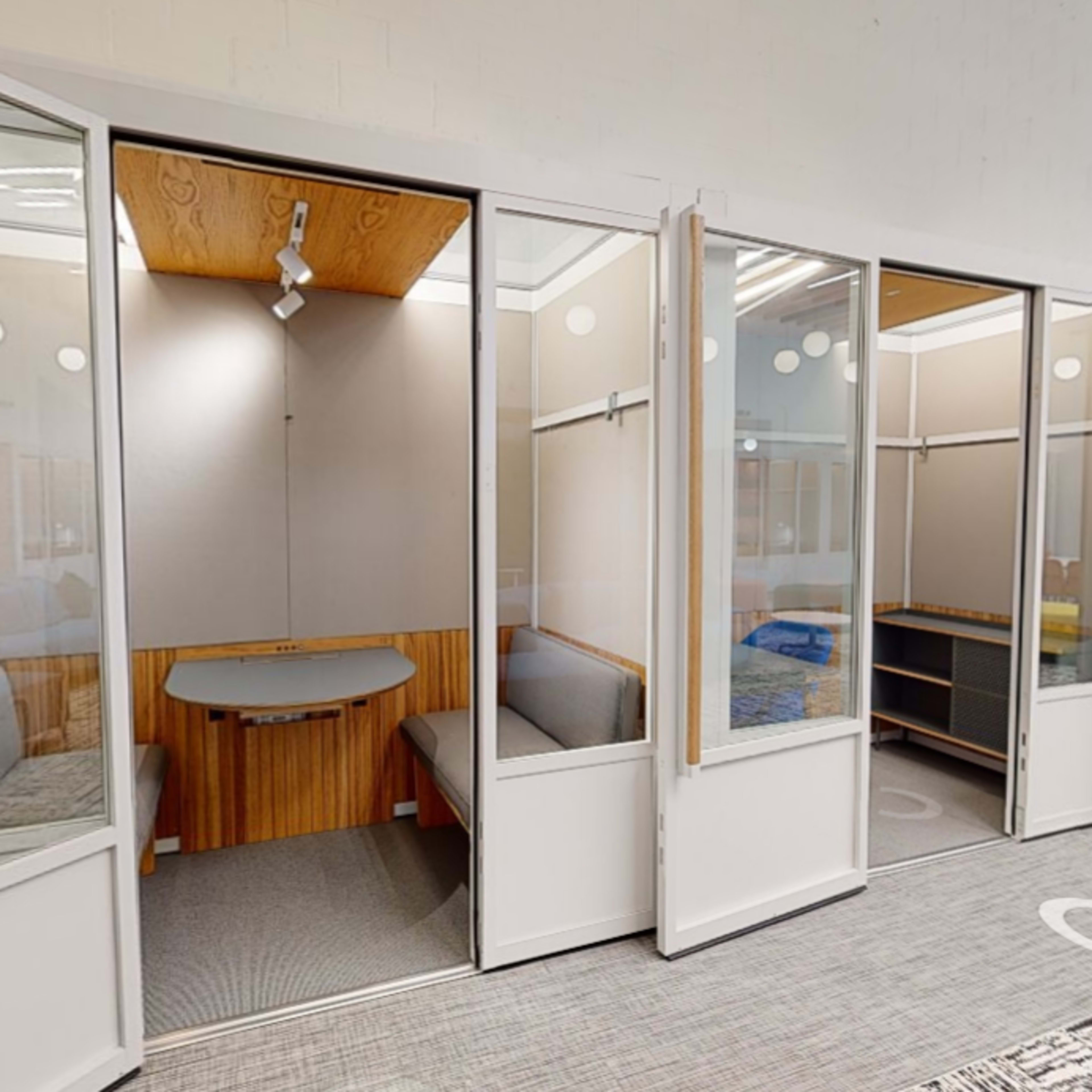 A row of glass-walled cubicles with wooden furnishings and seating areas in a modern office space.