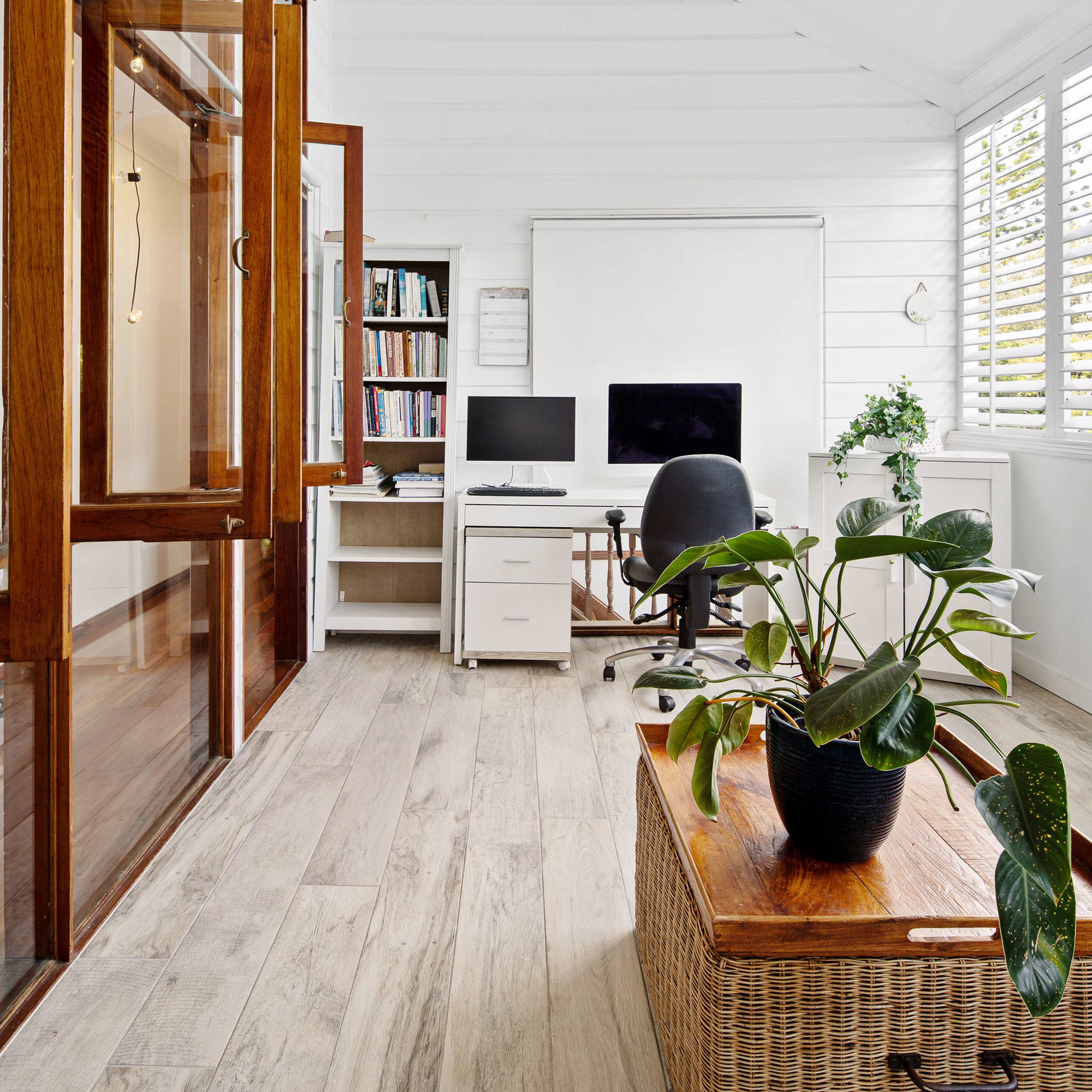 A home office with a wooden desk, a computer setup, a shelf filled with books, and a potted plant on a wicker storage chest.