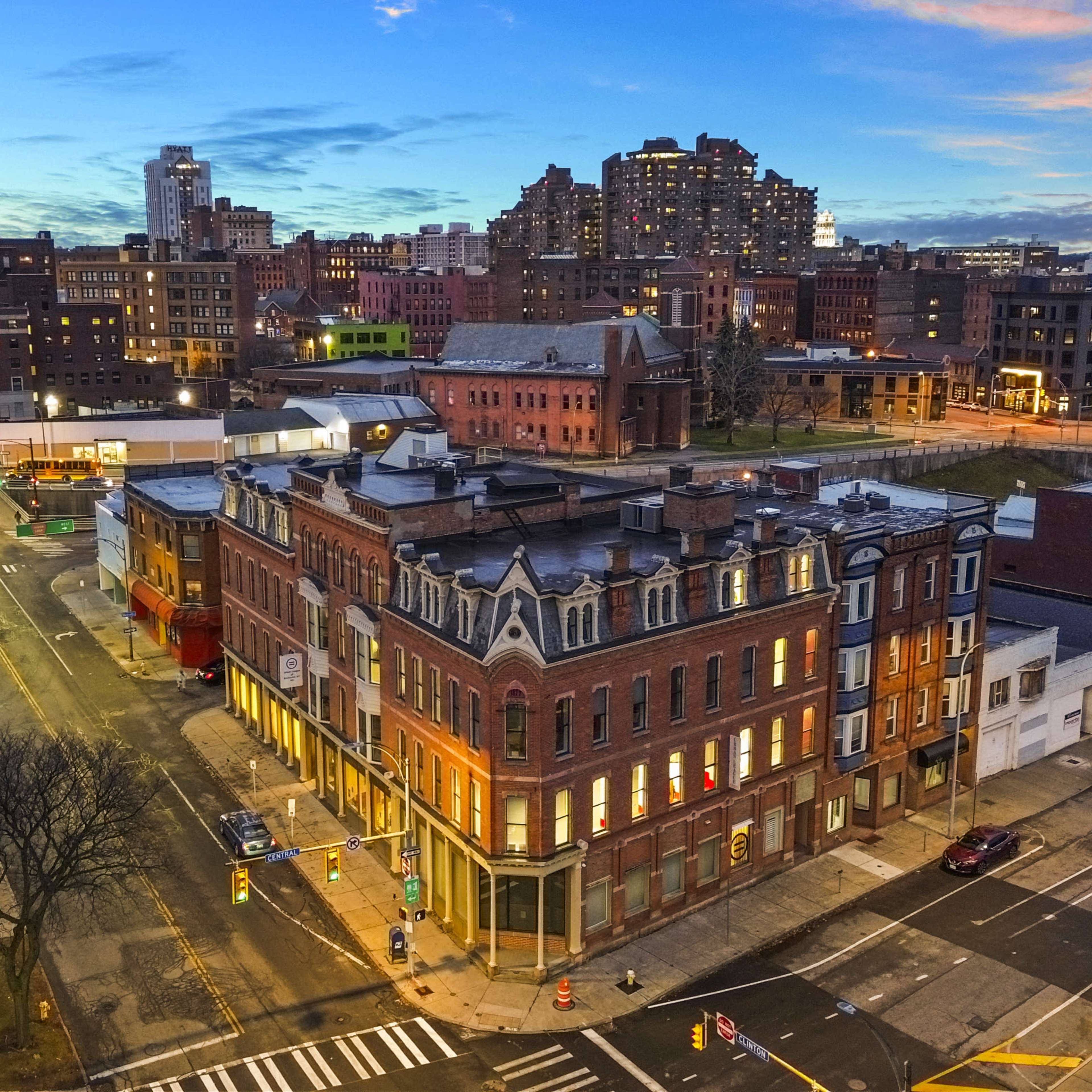A vibrant cityscape at dusk, featuring a historic building at a street intersection with modern skyscrapers in the background.