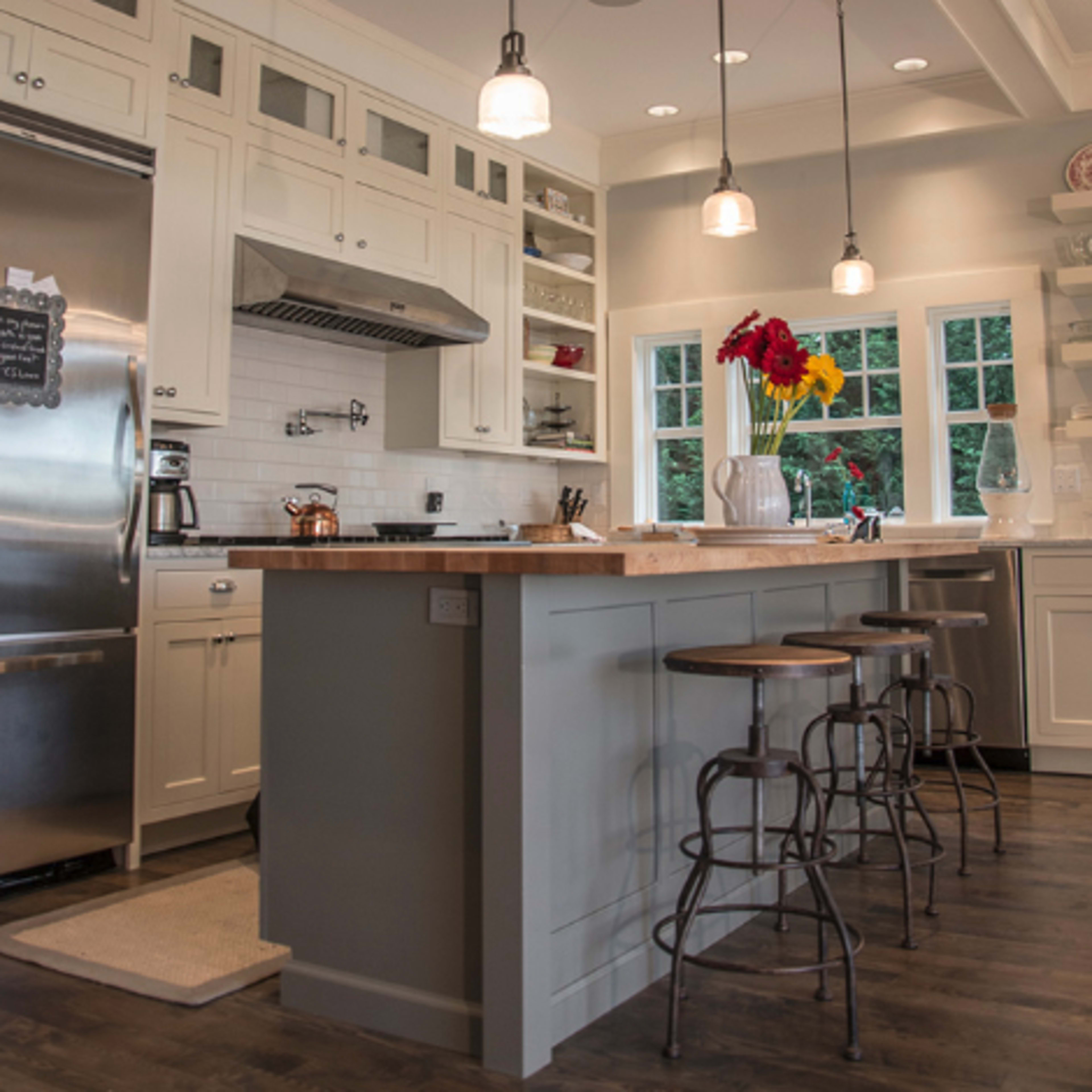 A modern kitchen features white cabinetry, a stainless steel refrigerator, and a central island with three metal stools beneath pendant lighting.