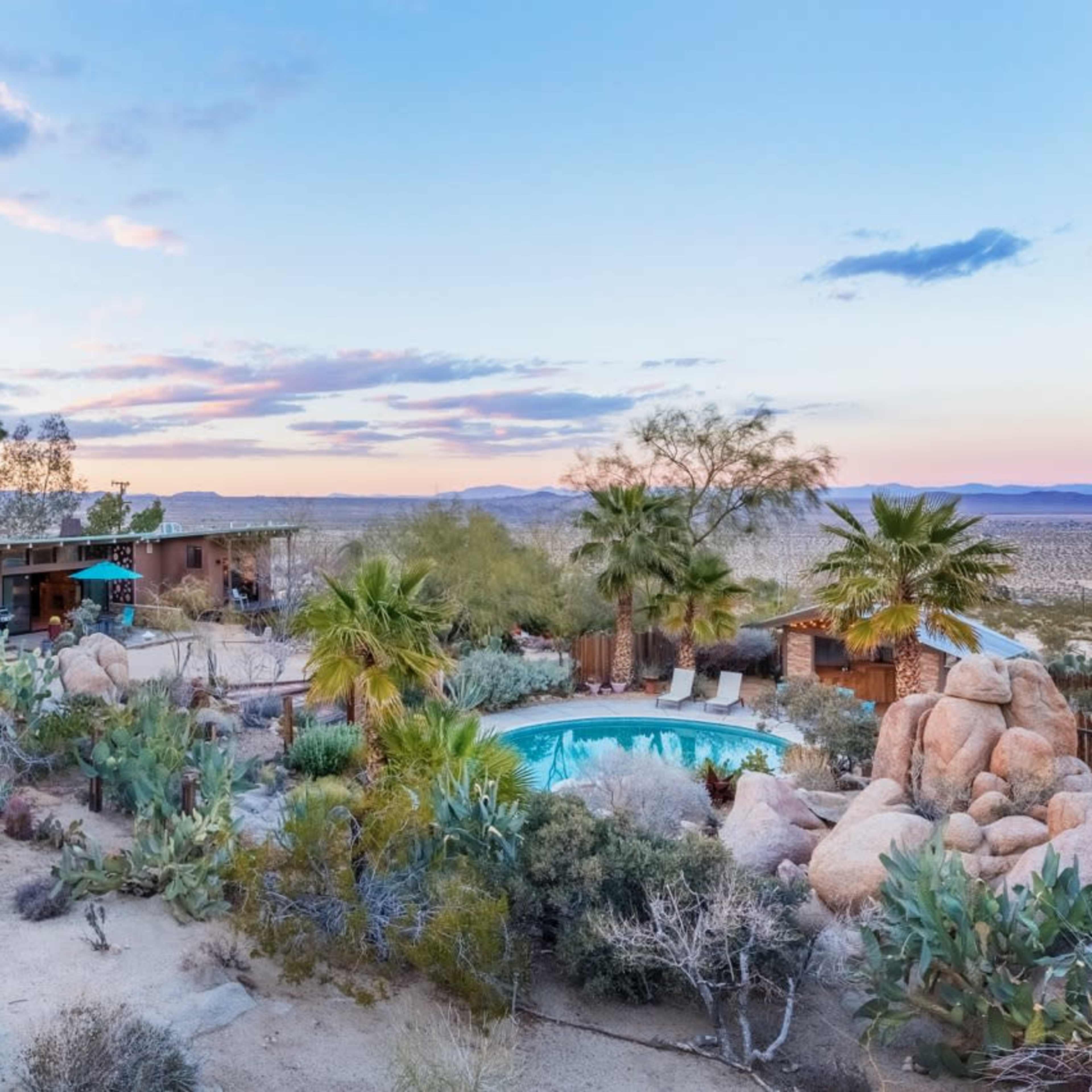 A desert landscape featuring a modern house with a swimming pool surrounded by large rocks and desert vegetation.