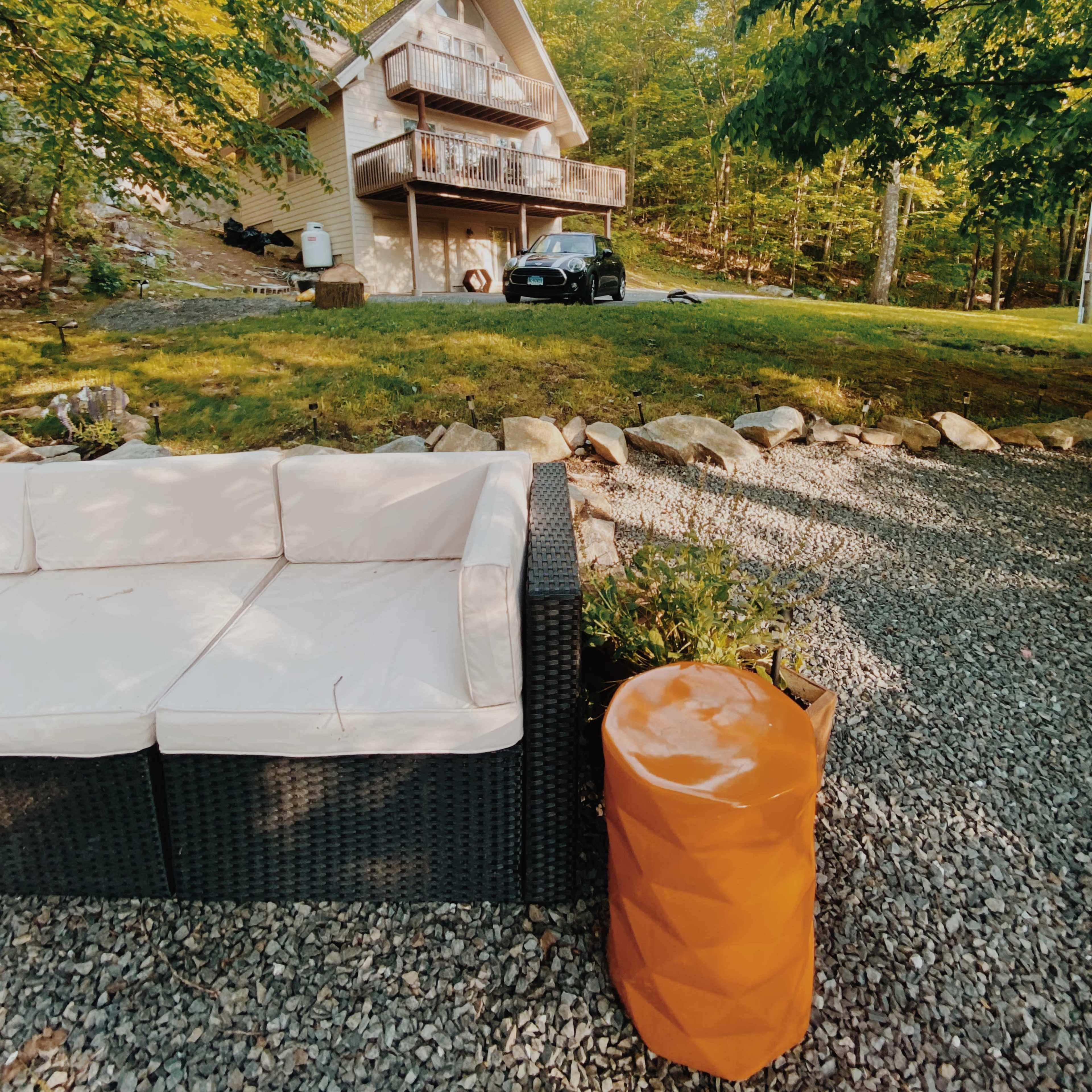 A two-story wooden house is situated on a grassy area surrounded by trees, with a modern couch and an orange side table in the foreground on a gravel surface.