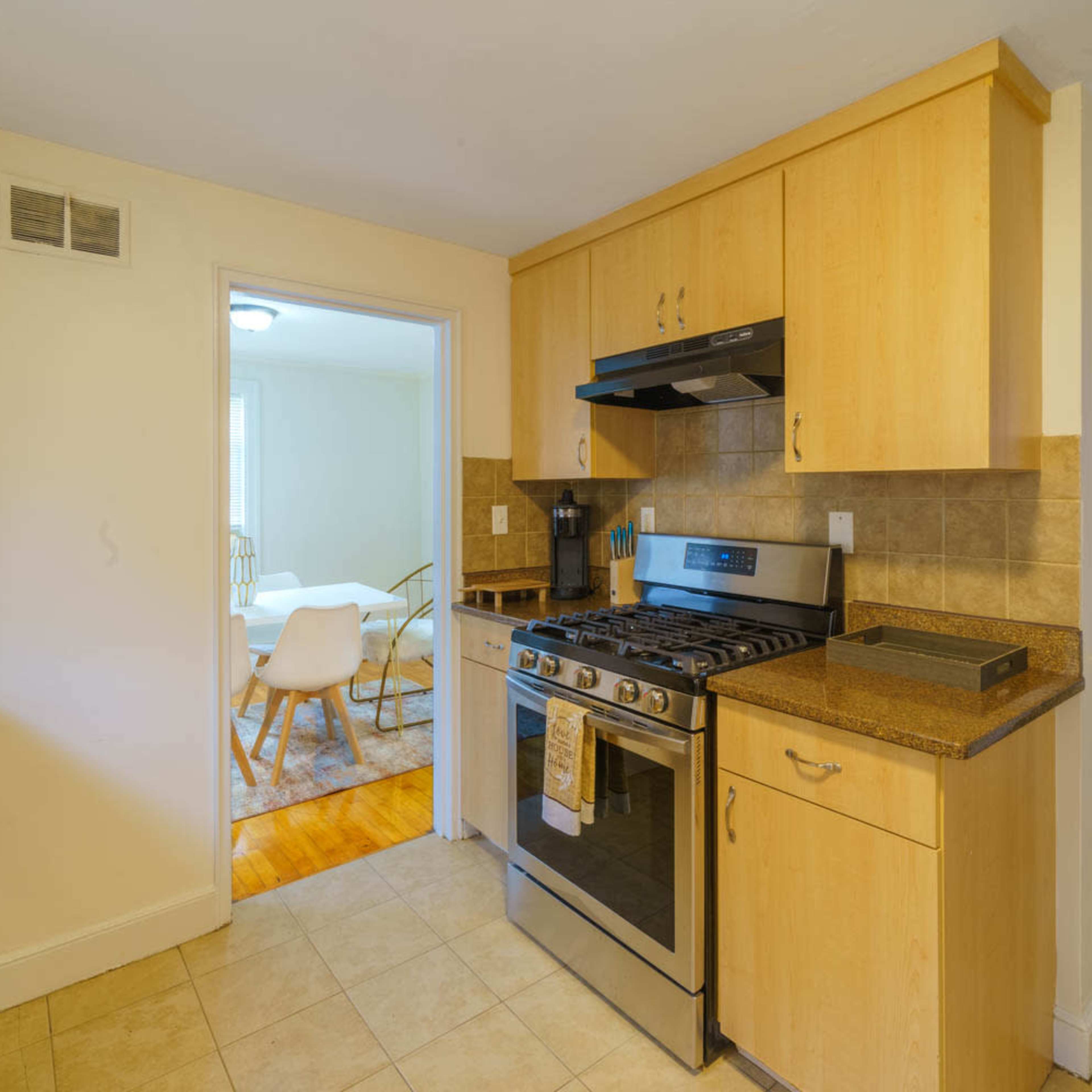 A modern kitchen features wood cabinetry, a gas stove, and tiled flooring, with a doorway leading to a dining area in the background.